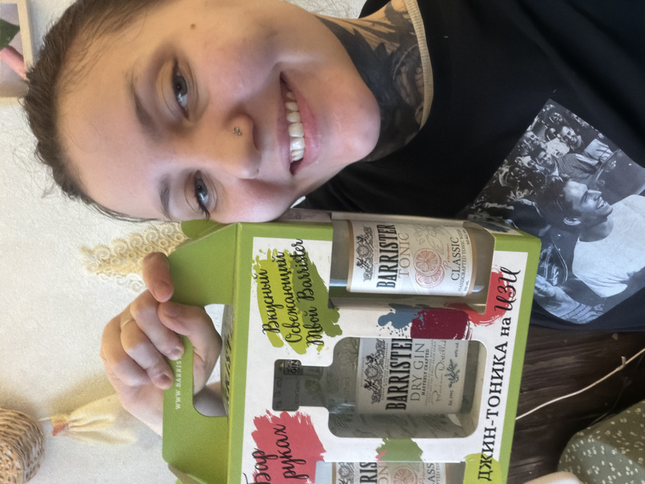 A woman smiles, holding a green box containing Barrister gin and tonic. The box features Russian text and the words "Barrister Dry Gin" and "Barrister Tonic Classic" in English. The woman has a nose piercing and tattoos on her neck. She wears a black t-shirt with a black and white image of a man on it. A decorative floral item rests on a surface behind the box. The text on the box translates to "Delicious Refreshing Your Barrister," "Bar in hands," and "Gin-tonic on easy".