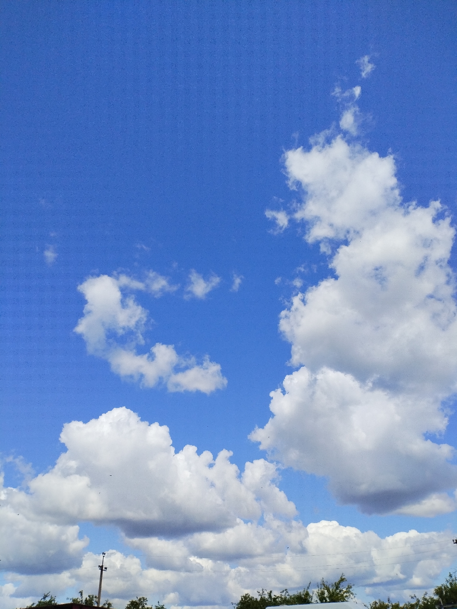 The image depicts a bright, sunny day with a vibrant blue sky filled with scattered white cumulus clouds. The lower portion of the image shows a tree line with foliage, suggesting an outdoor, possibly rural or suburban setting. A utility pole with wires is visible in the middle ground, indicating human infrastructure in the environment. The overall scene conveys a peaceful and serene atmosphere typical of a clear day. The location of Вароніцкі сельскі Савет, Belarus is noted as the setting.