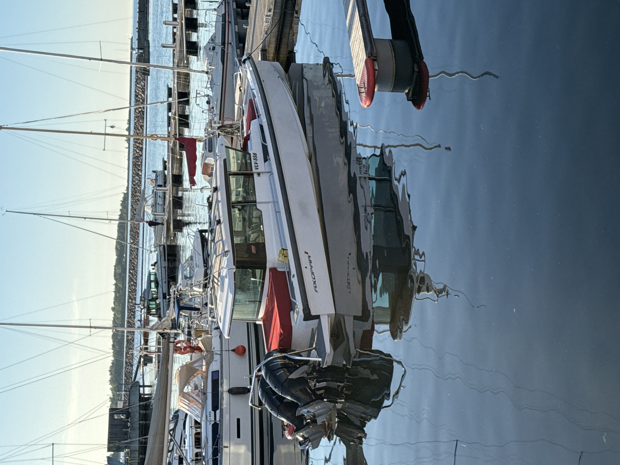 This image depicts a marina in Tallinn, Estonia, under clear daylight conditions. The foreground and mid-ground are dominated by calm, dark blue water exhibiting detailed reflections of vessels and their rigging. A white Axopar powerboat, marked with "AXOPAR" on its starboard side and the registration "VLV-558," is centrally positioned, featuring twin black outboard engines and red aft deck cushions. To its immediate left, a white sailing yacht with a blue hull stripe and a tan canvas dodger displays the registration "VLJ-669." Numerous additional sailboats and powerboats are moored at various wooden and concrete floating docks throughout the marina. One person is observed standing on a dock in the mid-ground, near a sailboat. The background features a stone breakwater, behind which a distant treeline and industrial structures are visible beneath a cloudless, light blue sky. A red circular fender is attached to a dock structure in the lower right.