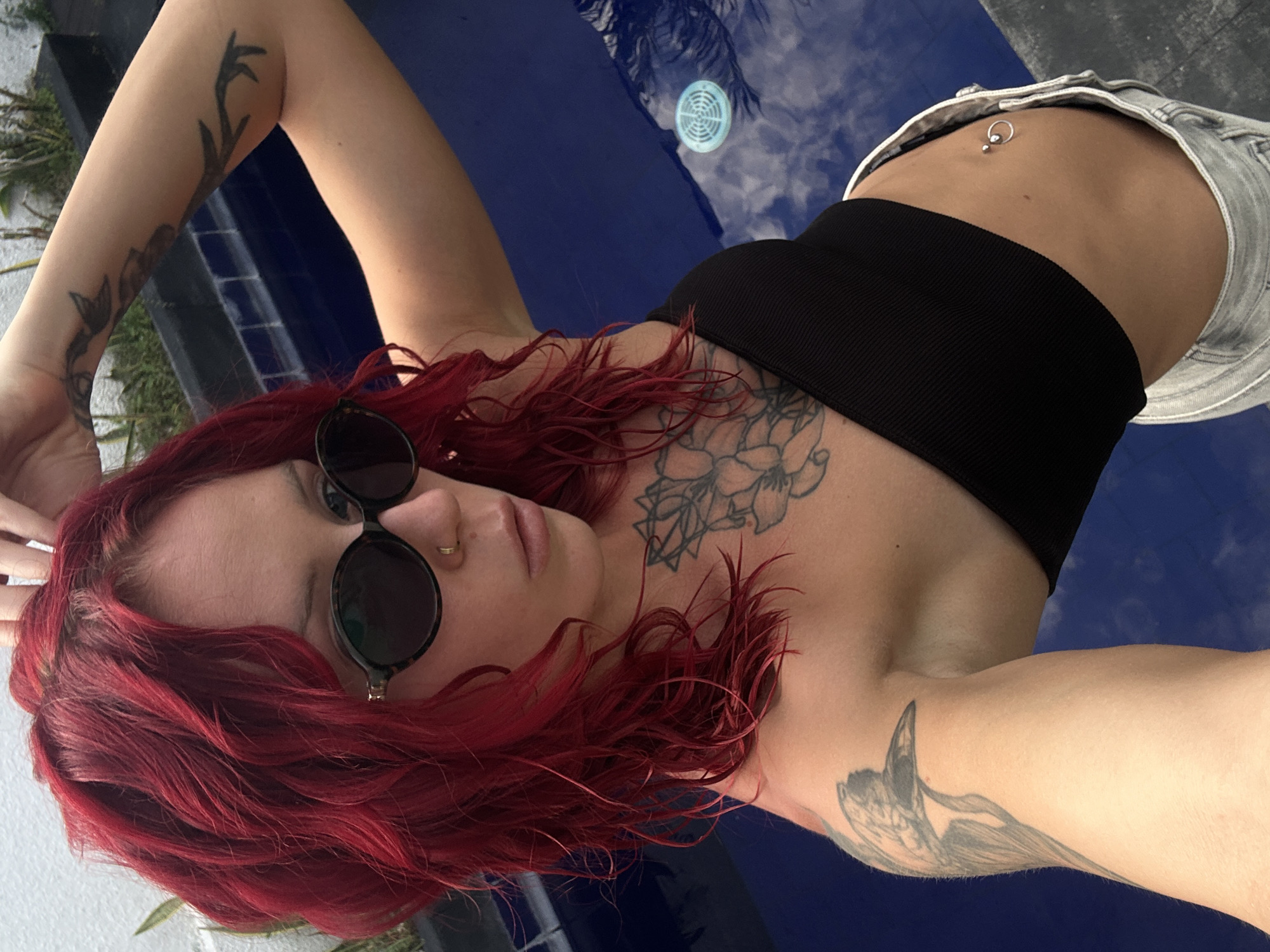 A young woman with red hair is lying down outdoors. She is wearing a black crop top, light-wash denim shorts, and round sunglasses. She has tattoos on her arms and chest, and a belly button piercing. She appears to be next to a blue pool or water feature. The location is outdoors in Dalung, Indonesia.