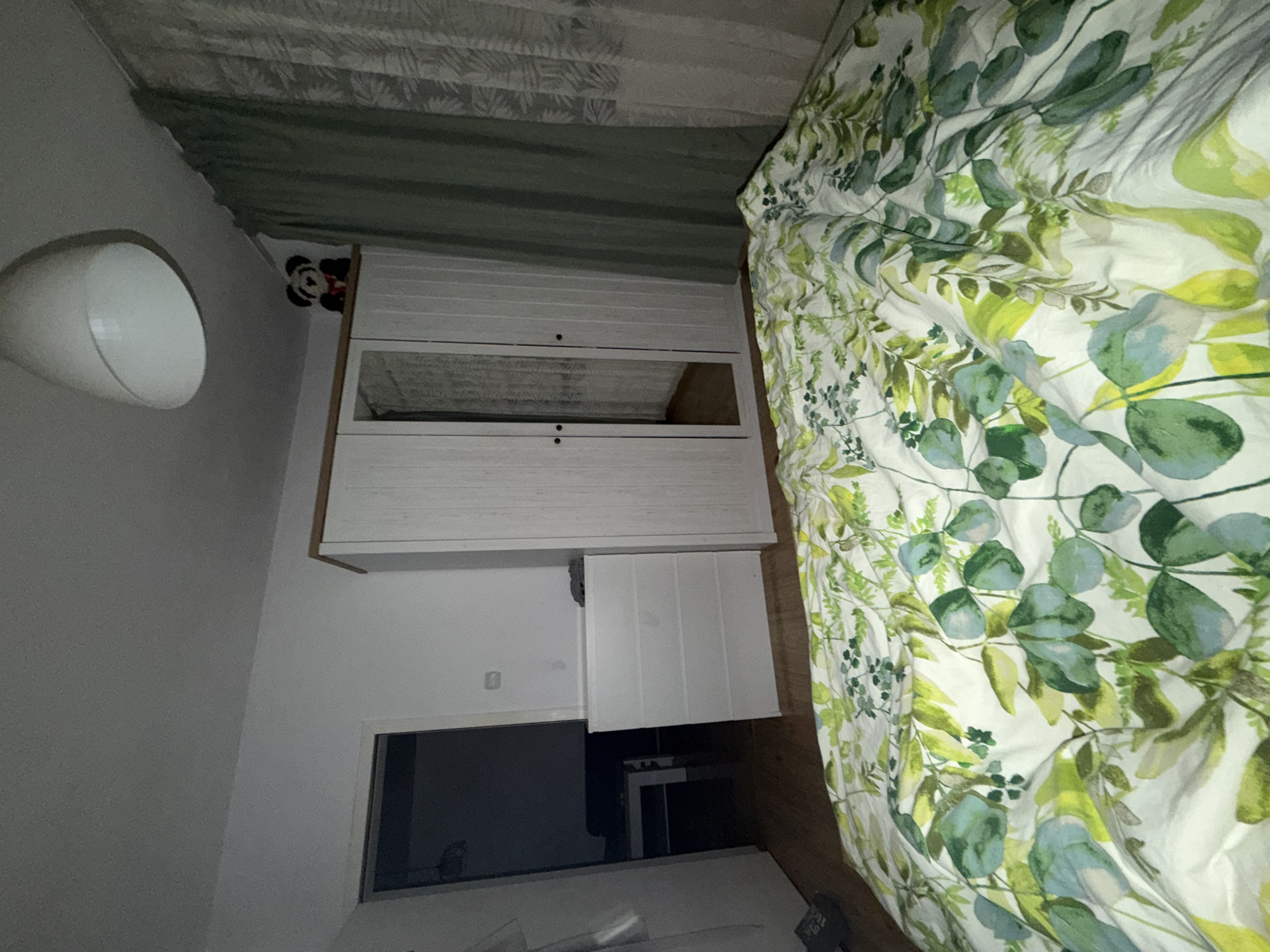 The image shows a bedroom in Plovdiv, Bulgaria. The room features a white wardrobe with mirrored doors, a chest of drawers, a white ceiling lamp, and green curtains. A stuffed teddy bear is perched on top of the wardrobe. The bed is covered with a duvet featuring a green leaf pattern. The floor is made of wooden planks. A doorway leads to another room, which appears to be a living area with a sofa and coffee table. The lighting in the room is dim, suggesting it is nighttime or the curtains are drawn. There are no people visible in the image.