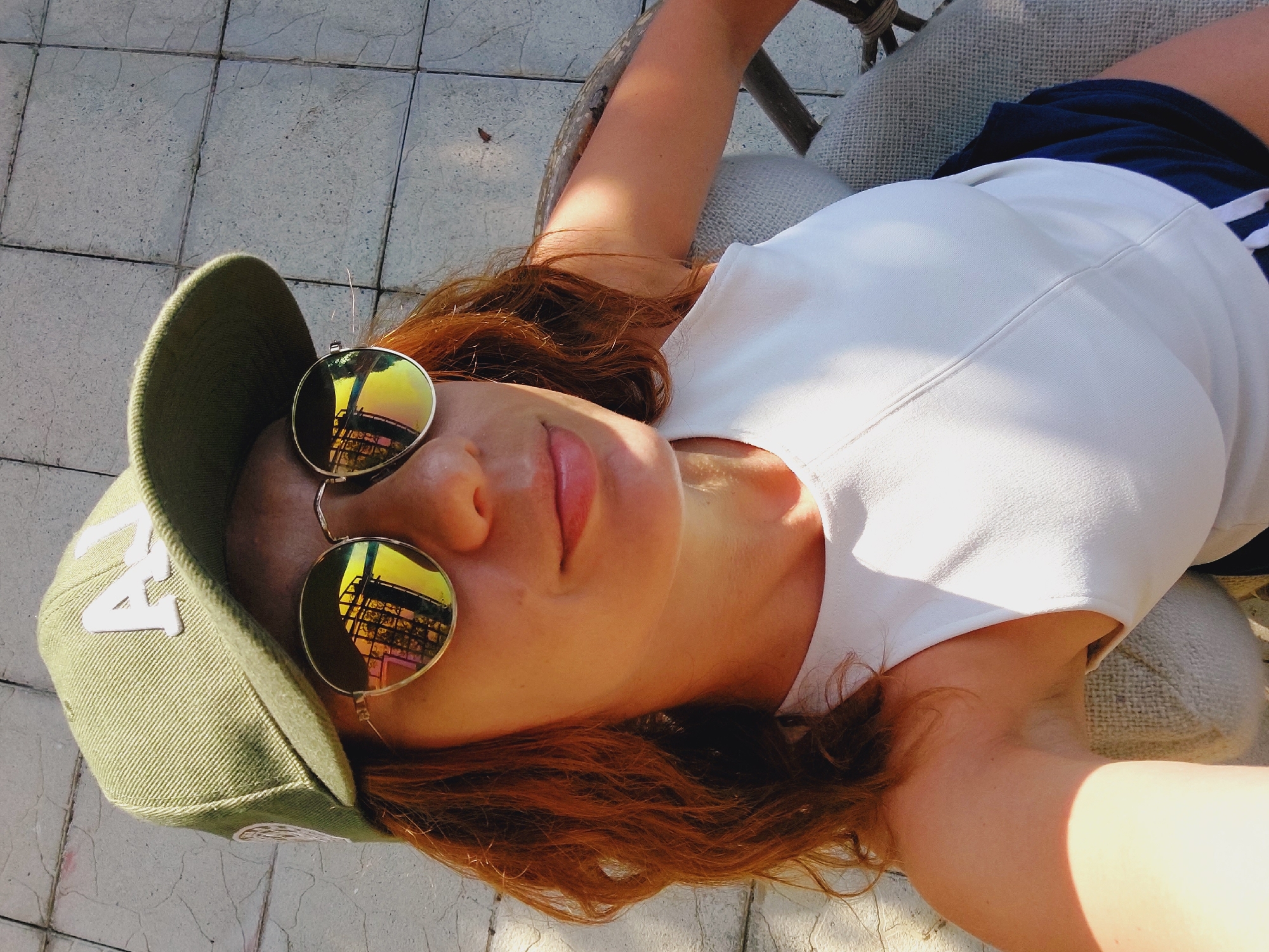 A medium shot, taken from a high angle, shows a woman with auburn hair wearing a green baseball cap, mirrored sunglasses, and a white sleeveless top. The sunglasses reflect a building with yellow and pink panes. She is seated outdoors on a chair with a textured, grey seat, outdoors on a patio with stone tiles. The woman is looking at the camera and smiling. Her left arm is resting on the chair. The image appears to be a selfie taken on a sunny day. The specific location in the city of Sozopol, Bulgaria, is not discernible from the visual evidence.