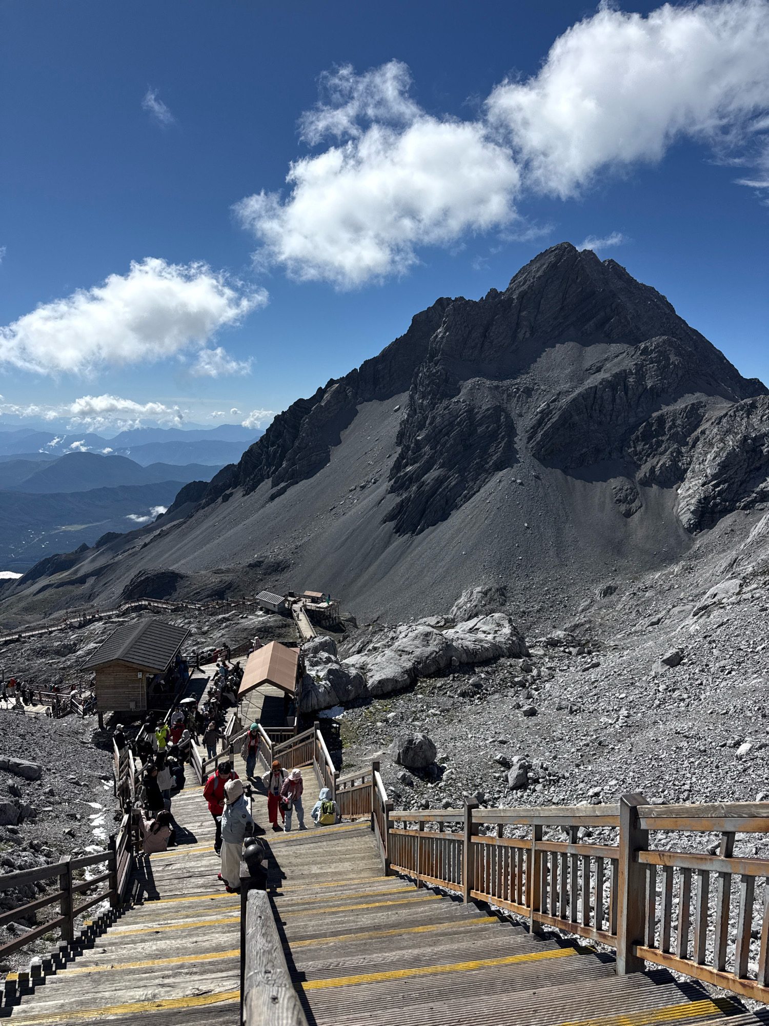 The image depicts a high-altitude mountainous scene in Yulong Nakhi Autonomous County, China, likely Jade Dragon Snow Mountain. A prominent, rugged, dark gray mountain peak dominates the upper portion of the frame, its slopes covered in scree and rock. Below the mountain, a winding wooden boardwalk with railings guides a stream of people. Several small wooden structures, possibly rest stops or shops, are situated along the boardwalk. The sky is a vibrant blue with scattered white, fluffy clouds. In the distance, layers of hazy blue mountains recede into the background. People are seen walking, standing, and interacting on the boardwalk, dressed in casual clothing, with some carrying backpacks or wearing hats. The overall impression is of a tourist destination offering scenic mountain views and accessible pathways.