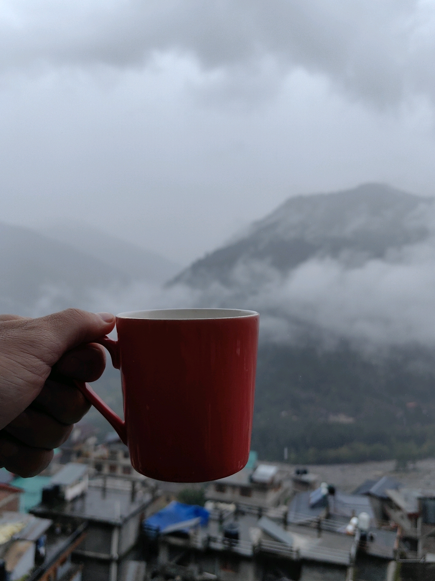 A hand is holding a red coffee mug against a misty mountain landscape and buildings. The mug is positioned in the foreground, obscuring some of the background details. The mountains are shrouded in fog, with only the darker shapes of their peaks visible. Below the mountains, there are several buildings, likely houses or shops, with some roofs covered in blue tarpaulin. The overall atmosphere is damp and overcast, suggesting a cloudy or rainy day in the city of Vashisht, India.