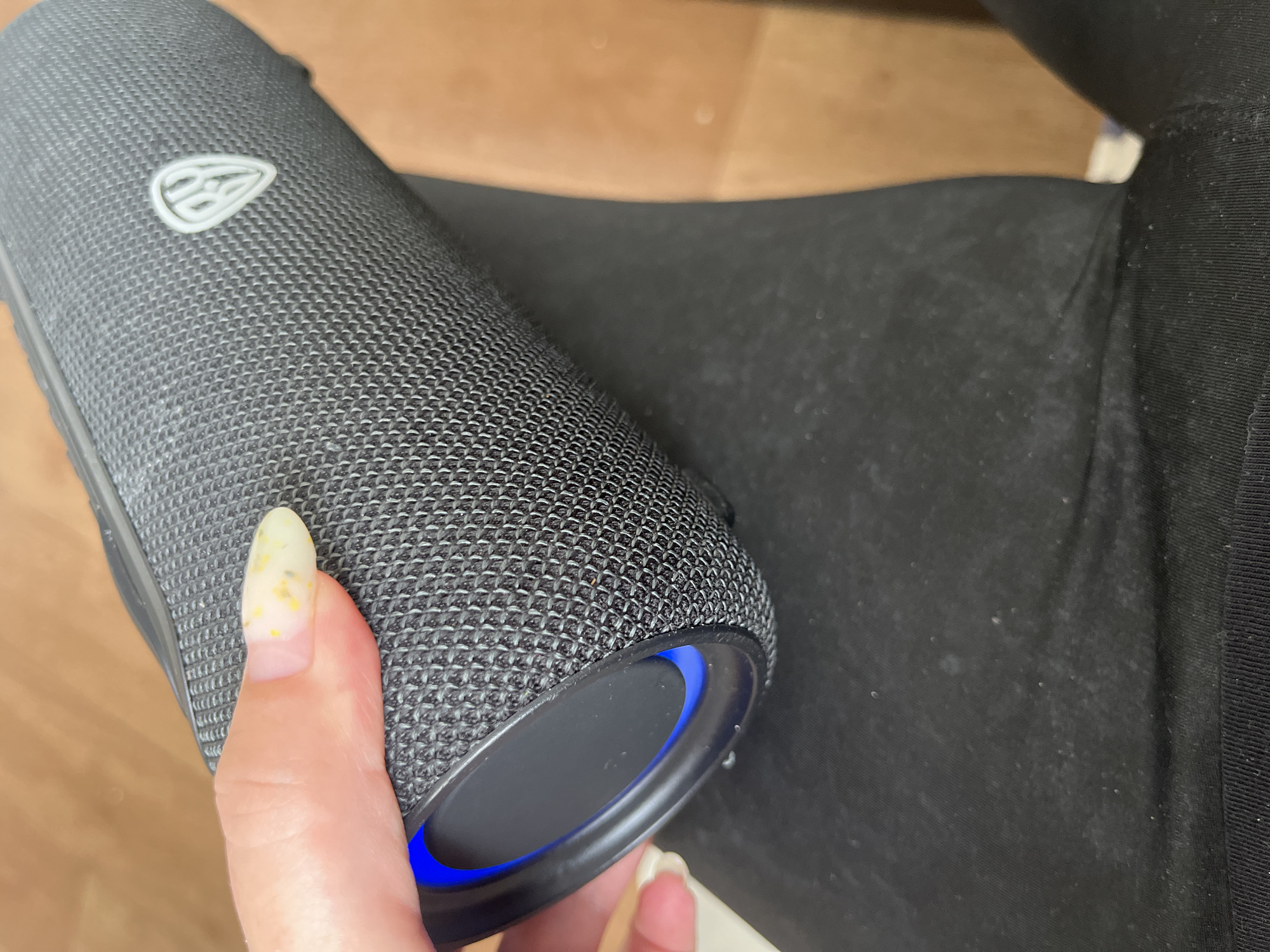 The image shows a close-up, overhead view of a person's hand holding a black, cylindrical Bluetooth speaker. The speaker has a textured surface and a blue LED light visible on one end. The person's fingernail is decorated with a yellowish and speckled design. The speaker is resting on black fabric, which appears to be clothing. The background consists of a blurred brown surface, possibly a floor or table. The image does not contain any information that can be used to determine its location in the city of Chelyabinsk, Russia.
