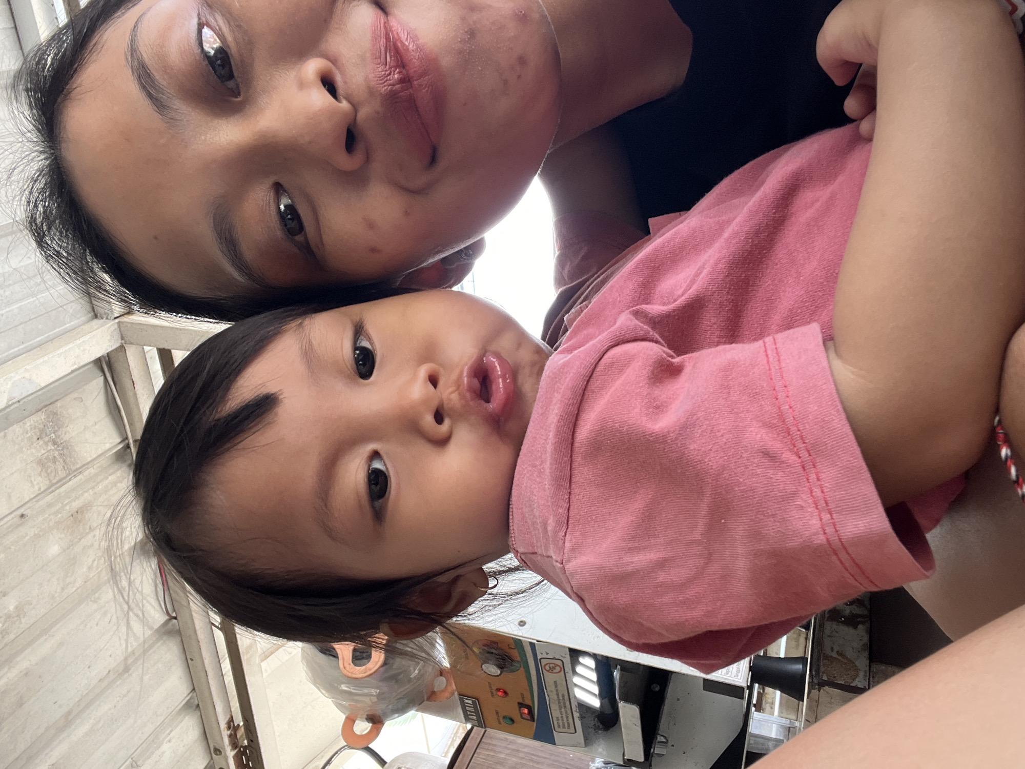A close-up selfie captures a woman and a child in what appears to be an outdoor or semi-outdoor environment, possibly a covered area judging by the corrugated metal ceiling visible in the upper portion of the image. The woman is on the right, her face filling much of the frame, with dark hair pulled back. She is smiling, and her lips are painted with a muted pink lipstick. The child is positioned to the left of the woman, looking towards the camera with wide eyes and puckered lips. The child is wearing a pink t-shirt. In the background, partially obscured, is a piece of equipment that looks like a sealing machine, with branding that includes the word "MATRIX." The setting suggests a commercial or workshop environment within Kecamatan Pati, Indonesia.