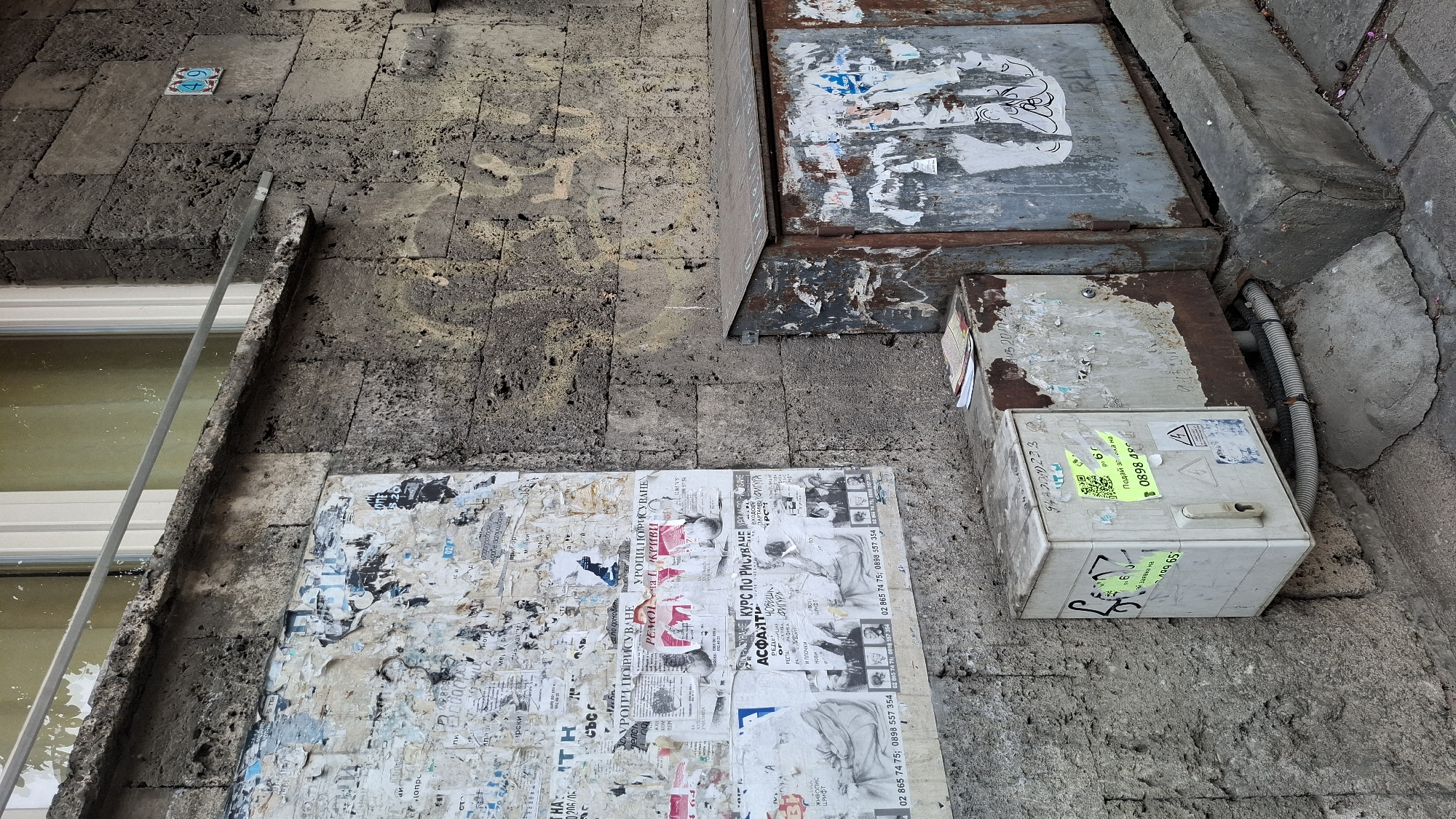 This is a medium shot, taken from a high angle, of an exterior wall in Sofia, Bulgaria. The wall is constructed of rough, weathered stone blocks. The scene is cluttered with various objects and aged posters.
Dominating the lower half of the image is a large, tattered bulletin board covered in numerous layers of ripped and peeling posters, primarily black and white newspapers or advertisements. Some posters display images of people, including portraits and group shots. Visible text in Cyrillic includes phrases like "КУРС ПО РИСУВАНЕ" (drawing course), "ЖИВОПИС" (painting), "АСФАЛТИ" (asphalt), and phone numbers.
To the right of the bulletin board, a light-colored electrical or utility box is visible, attached to a rusted metal casing. This box has several stickers on it, including one with a QR code, and another with Cyrillic text and a phone number. A gray corrugated pipe extends from the rusted casing into a concrete drain.
Above the bulletin board and to the right, a large, rusted metal container or bin is present, also covered in torn posters and graffiti.
The overall impression is one of urban decay and neglect. The lighting suggests it is daytime, with no strong shadows indicating the time of day. The weather appears to be dry.