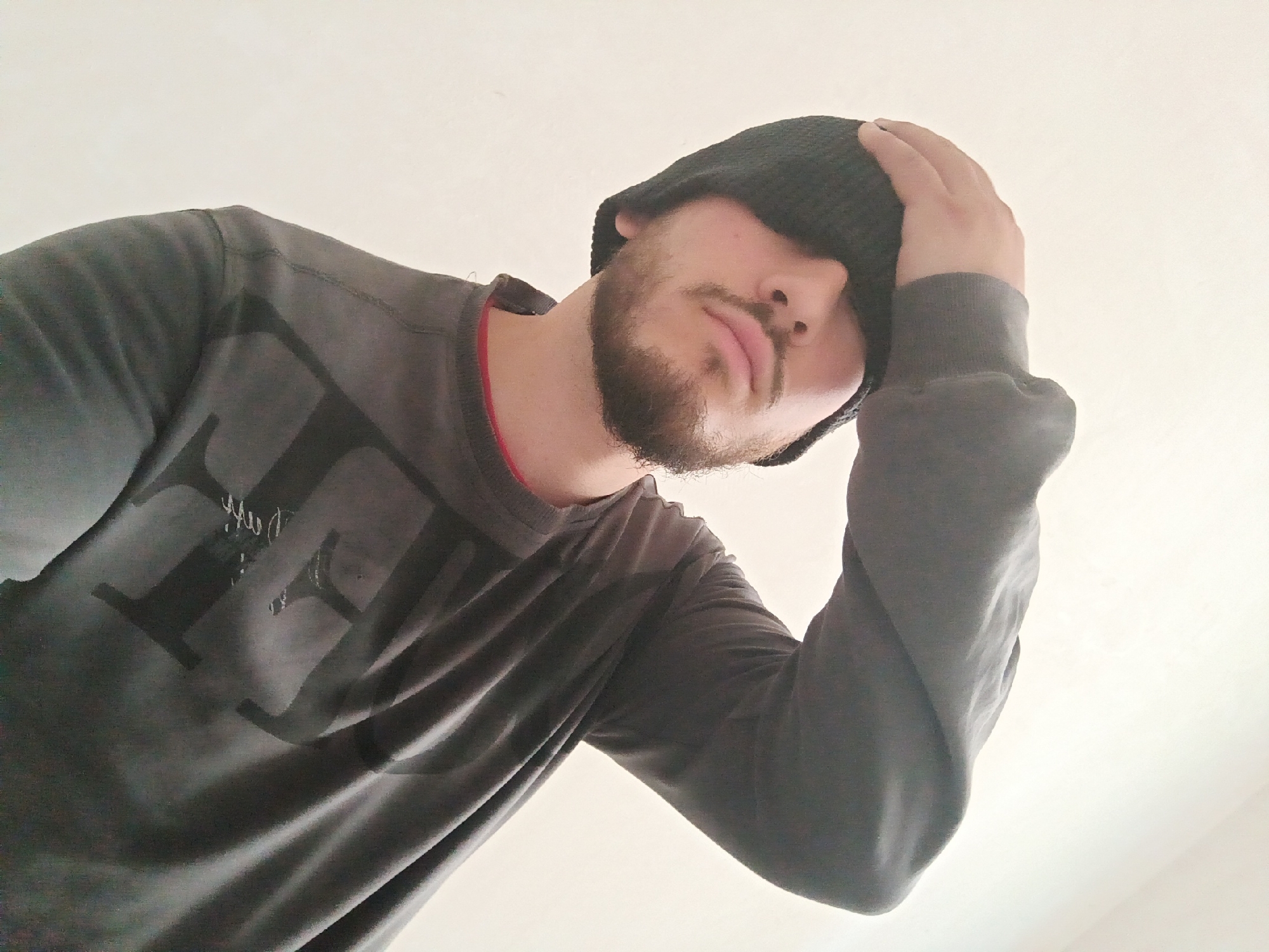 A male individual is depicted from a low-angle perspective against a plain, light-colored background. He has a light brown beard and mustache. He wears a dark grey long-sleeved sweatshirt with a subtle dark graphic on the chest, and a red trim is visible around the collar. A black knit beanie is pulled down, completely obscuring his eyes. His right hand is raised, with fingers splayed, resting on the top of the beanie. His head is tilted slightly to his right, and his lips are parted in a neutral expression. No specific location within the city of Sofia, Bulgaria, can be determined from the visual information provided. The environment appears to be an indoor setting with a nondescript wall or ceiling. No interactions are visible.