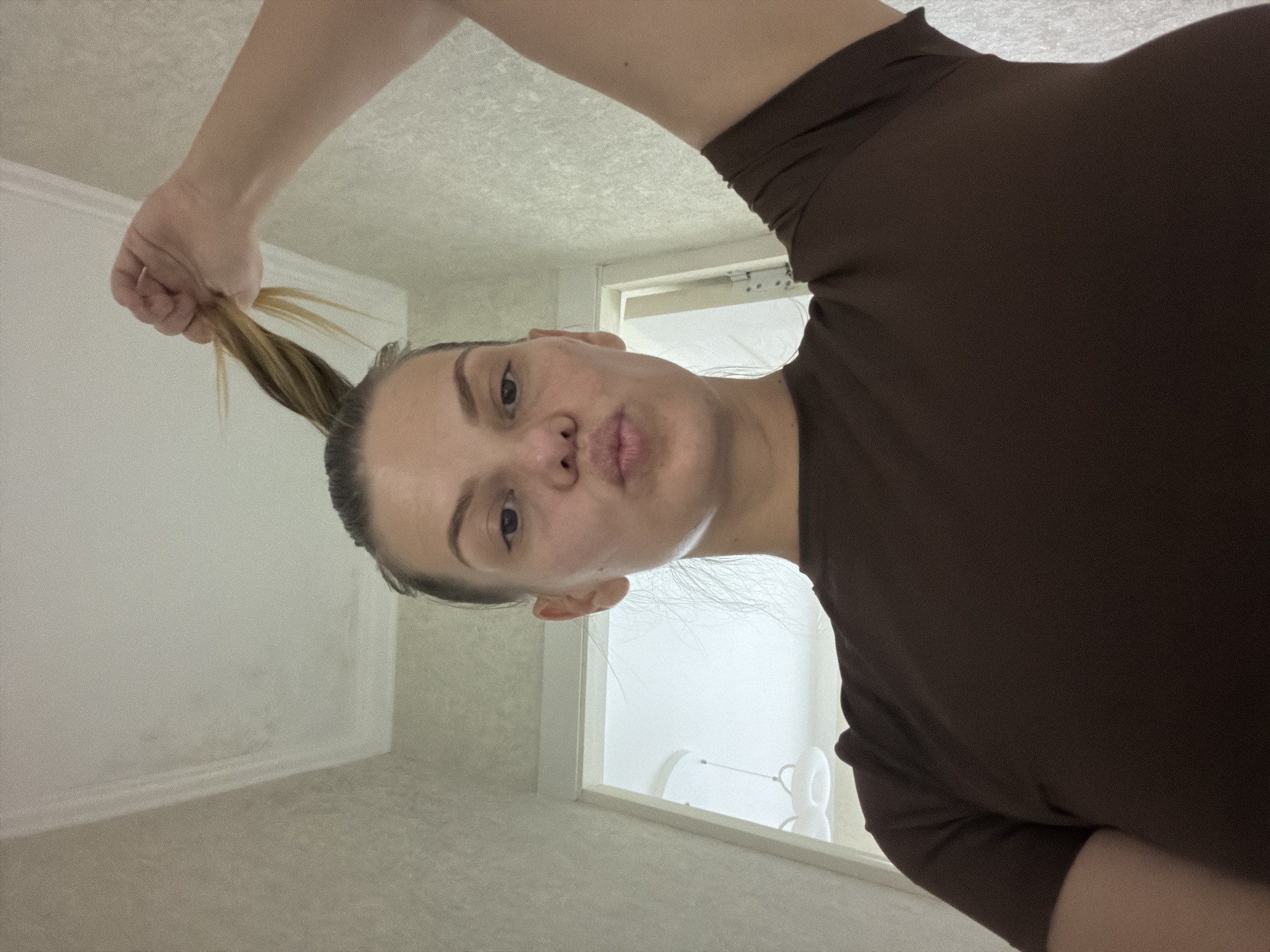 A woman is holding her ponytail and making a duck face in a selfie. She is indoors, in what appears to be a hallway or entryway of a home. The walls are textured and light-colored, and the ceiling has a white textured tile pattern with a decorative molding. A doorway is visible in the background, leading to a brightly lit room with a modern light fixture. She is wearing a plain brown t-shirt. The image was taken in Chirchik, Uzbekistan.