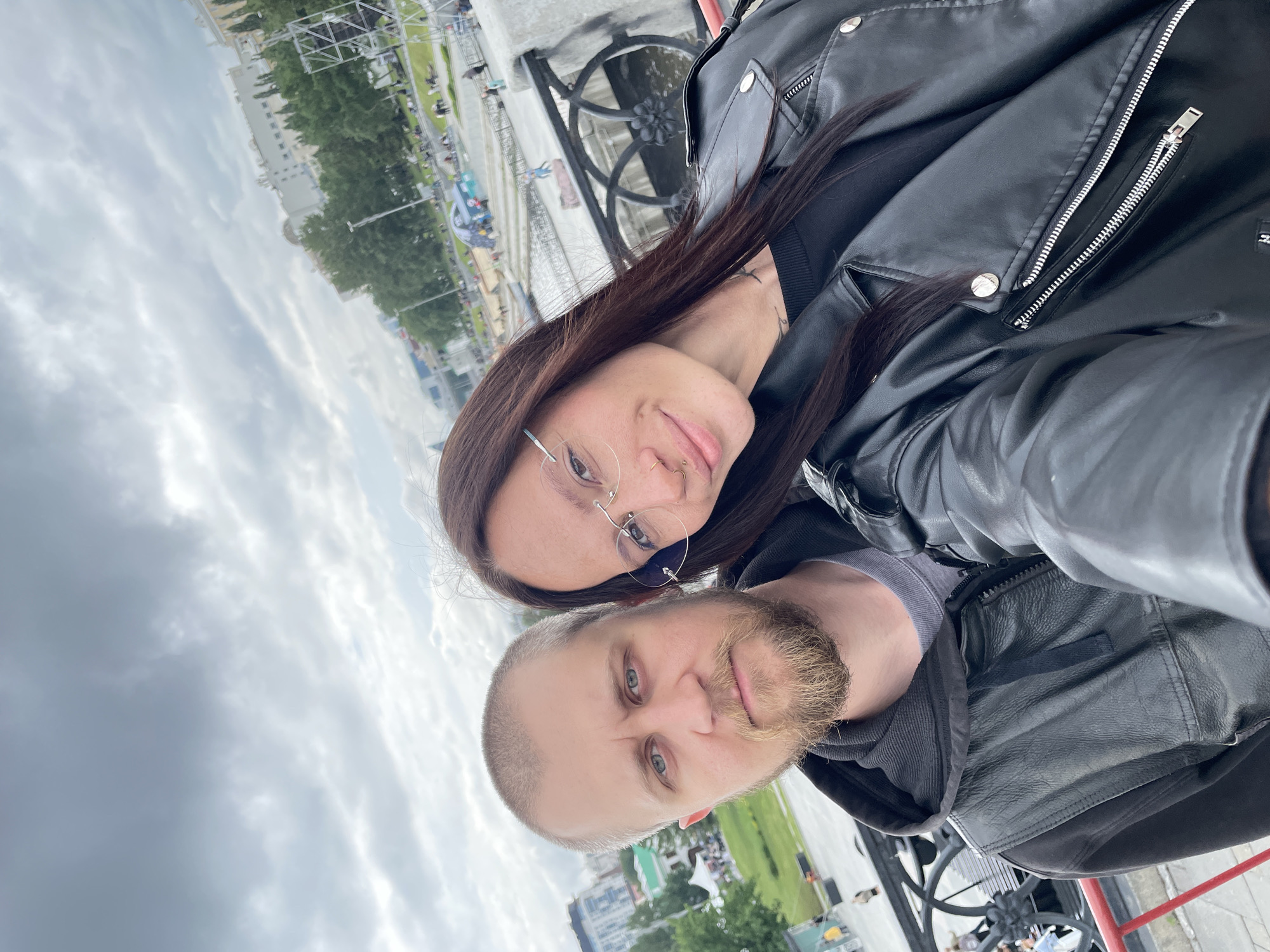 A selfie-style portrait features two individuals, a man and a woman, positioned in the foreground, looking directly at the camera. The setting is an urban outdoor environment, identifiable as the Iset River Embankment in Yekaterinburg, Russia, specifically near the Plotinka dam or Historical Square.
The man, on the left, has light skin, blue eyes, a short beard, and a closely cropped hairline. He wears a grey t-shirt under a dark grey hooded sweatshirt, over which he wears a black leather jacket. His expression is neutral.
The woman, on the right, has long, dark brown hair, light skin, and wears rimless eyeglasses. She has a nose piercing on her right nostril and a small tattoo visible on her neck/collarbone area. She wears a black top under a black leather biker jacket with silver hardware. Her lips are slightly parted in a subtle smile.
Behind the individuals, a decorative black metal railing with circular motifs, characteristic of the Iset River embankment, is visible. Beyond the railing, a body of water (the Iset River) flows to the right. On the far bank, a promenade or park area is visible, with numerous people strolling and several tents or structures, including a prominent blue tent. Trees line the embankment. In the mid-ground and background, various multi-story urban buildings, consistent with Yekaterinburg's architecture, rise under an overcast sky filled with grey clouds. The lighting suggests daylight, possibly late afternoon or early evening.
