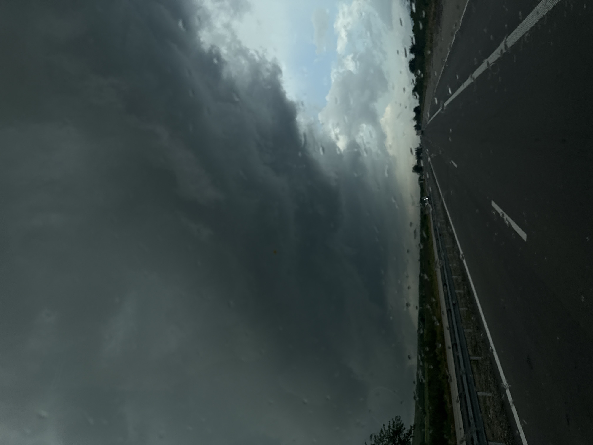 The view from a vehicle's windshield reveals a dark, stormy sky dominating the left side of the frame. Raindrops are visible on the glass. To the right, a section of asphalt highway stretches into the distance with a painted white line marking lanes. A strip of greenery borders the road. The location is identified as Zlatina, Bulgaria. No people or specific interactions are visible.
