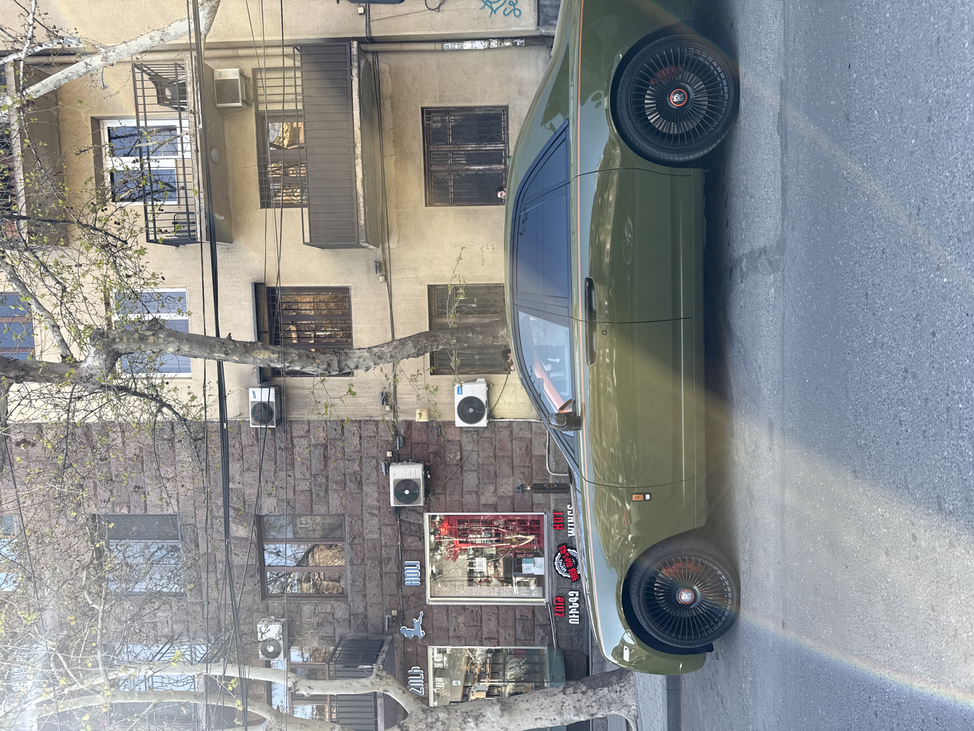 An olive green car, resembling a customized classic model, is parked alongside an urban street in Yerevan, Armenia. The vehicle features dark tinted windows, an orange or red interior, and distinctive multi-spoke black wheels with red center caps that bear a logo (possibly "RR"). A faint rainbow reflection stretches across the asphalt and the car's side, suggesting a sunny day with specific light conditions.
The car is positioned next to a row of multi-story buildings, characteristic of city architecture. The buildings display a mix of light, stucco-like facades and sections clad in darker, textured stone. Numerous air conditioning units are mounted on the exterior walls, and many windows are protected by black metal grates or feature small balconies. Electrical wires and cables are visible, running vertically down the building facades.
In the foreground, a deciduous tree with mostly bare branches and nascent green leaves partially obscures the view.
Notable details include:
- **Time of day:** Daytime, with bright sunshine casting reflections.
- **Location:** An urban street in Yerevan, Armenia, as indicated by the context and architectural style.
- **Visible text:** On the stone-clad building, a shop window features a red illuminated sign with "HOT" and a flame icon, along with "BURGER" (partially visible). Below this, a large sticker reads "GARAGE 66440" with additional Armenian script that appears to spell "GARAGE." To the left, other signs include fragmented Armenian text, one starting with "ՎԵՐ".