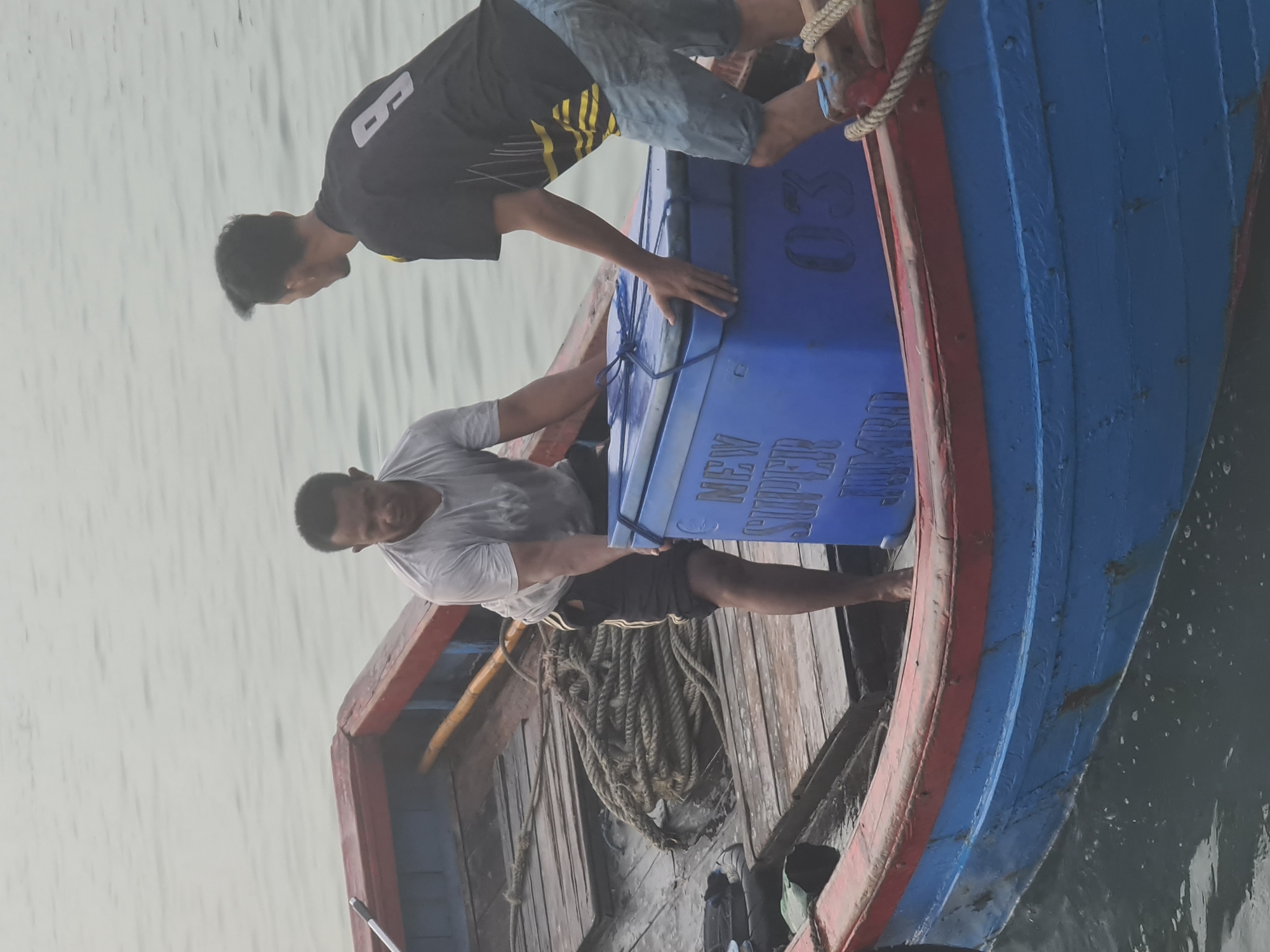 Two men are on a wooden boat in the water. The man on the left, wearing a grey t-shirt and black shorts, is bent over and holding a large blue cooler. The man on the right, wearing a black t-shirt with yellow stripes and denim shorts, is also holding the cooler. The cooler has "NEW SUPER JUMBO" written on it in white lettering, along with the number "03". The boat has wooden planks for flooring and a red and blue painted exterior. Ropes are coiled on the deck of the boat. The background is the water, which appears calm and grey. This scene is likely occurring in the waters of Lhokseumawe, Indonesia, a coastal city known for its fishing activities.