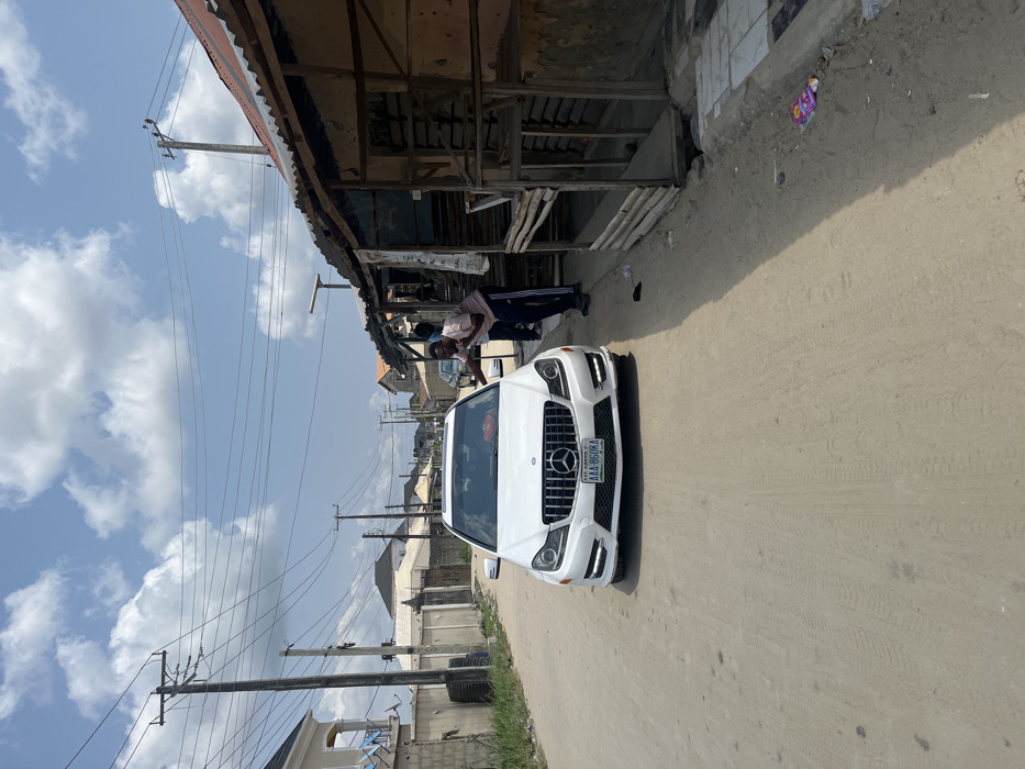 The image captures a street scene in Iranla, Nigeria, under a bright blue sky with scattered white cumulus clouds. A white Mercedes-Benz C-Class sedan, displaying a Lagos State license plate "AAA-860KA," is stationary on an unpaved dirt road, facing the viewer. A red object rests on its dashboard. To the immediate right of the vehicle, a male individual wearing a light pink short-sleeved polo shirt and dark track pants leans towards the passenger side of the car, with his right hand near the window frame and his left hand near his face, possibly gesturing or holding an object. A second male, partially obscured behind the first, stands further back near the car's rear passenger door. The right side of the road is bordered by a series of makeshift commercial structures or stalls constructed from wooden frames and topped with corrugated metal roofs, some exhibiting signs of rust and disrepair. Litter, including plastic wrappers, is visible on the dirt ground near these structures. The left side of the road features more developed residential buildings with concrete walls, one showing a light blue balcony railing, and perimeter walls topped with security elements. Multiple wooden utility poles with numerous overhead power lines line both sides of the street, extending into the background. A solar-powered street light is affixed to one of the utility poles on the right.