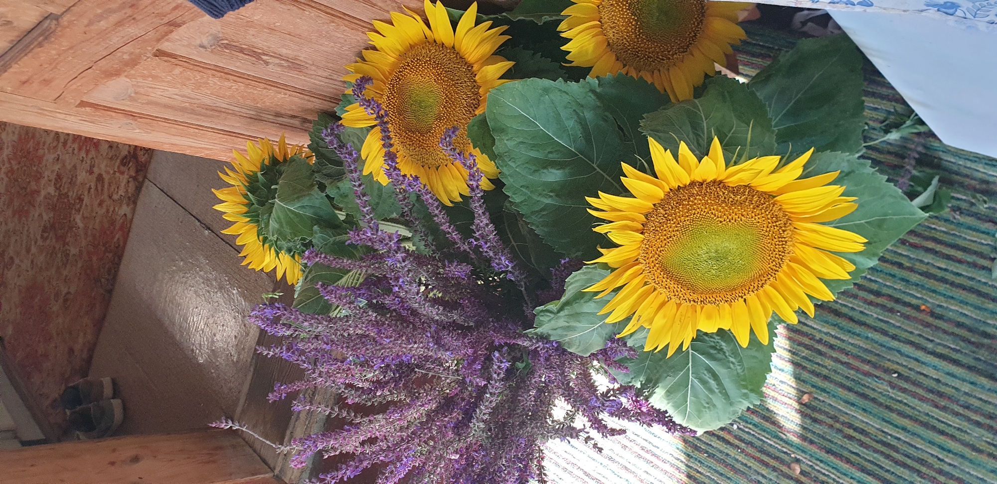 The image shows a close-up view of a bouquet of sunflowers and purple flowers, likely lavender, arranged near a wooden doorway. The flowers are placed on a striped rug, with a glimpse of shoes on a wooden floor in the background. The scene appears to be indoors, possibly in an entryway or porch area. There is no indication of the city of Kostanay, Kazakhstan, in the image.