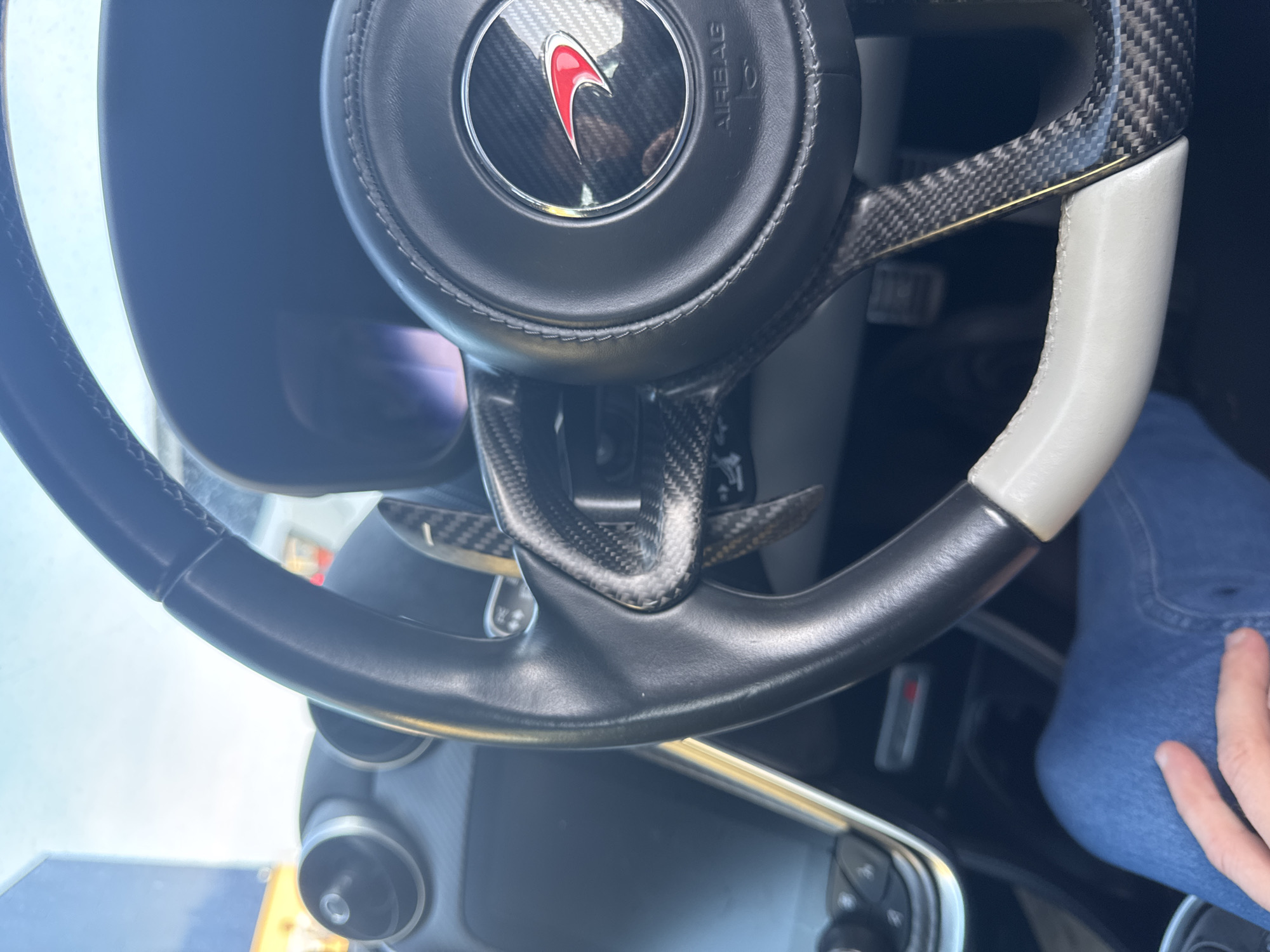 The image shows the interior of a vehicle, specifically the steering wheel. The steering wheel has a black leather grip, carbon fiber accents, and a white grip section. The central emblem is a red and silver logo within a black circle marked "AIRBAG." Paddle shifters are visible behind the wheel, constructed of carbon fiber. The dashboard is partially visible, including a control panel with buttons. A person's leg in blue jeans and hand are visible to the right. The city location Great Yarmouth and United Kingdom do not feature in this picture.