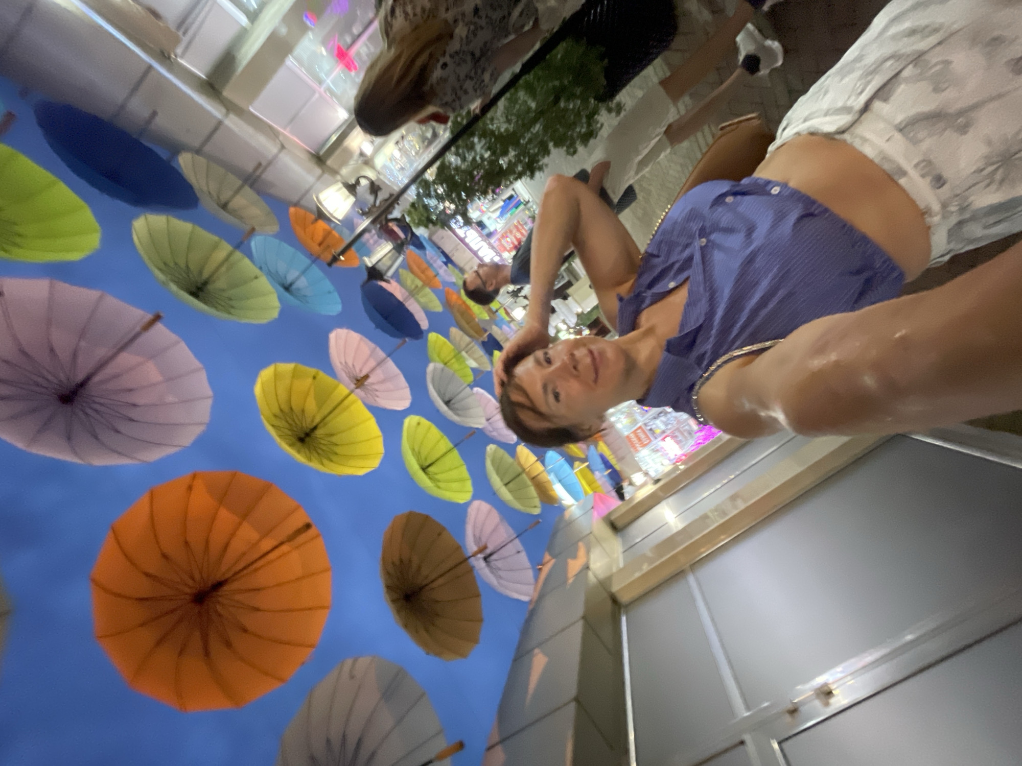 The image shows a street scene decorated with numerous colorful umbrellas suspended overhead. In the foreground, a woman is taking a selfie, wearing a blue and white striped crop top and white patterned pants. She has short brown hair and is smiling at the camera, with her arm extended. Behind her, a man in glasses and a dark t-shirt is visible, and further in the background, another woman with long brown hair is walking away from the camera. The street appears to be lined with shops, some of which have illuminated signs. The overall atmosphere suggests a lively urban environment, likely a pedestrianized area. This scene is located in Sochi, Russia, a resort city on the Black Sea coast, known for its vibrant atmosphere and summer decorations.