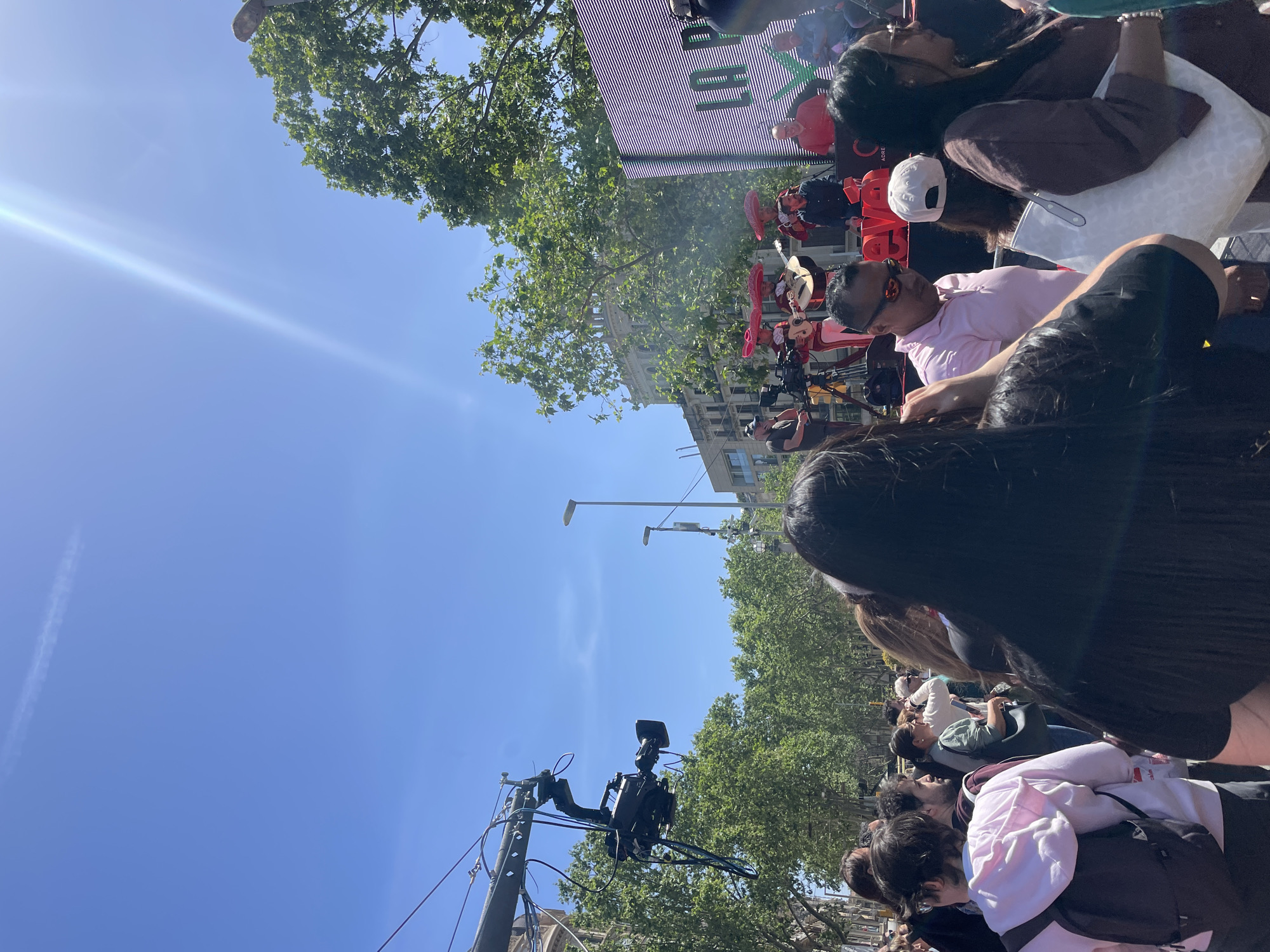 This image captures a vibrant outdoor cultural event taking place under a clear, bright blue sky in Barcelona, Spain. A strong sun flare is visible in the upper left, indicating a sunny day.
The scene features a stage area where performers, dressed in what appear to be traditional mariachi-style outfits complete with sombreros, are playing guitars. A large outdoor LED screen acts as a backdrop to the stage, displaying vertical scan lines and some partially visible green text, possibly reading "H" and "LL". Below this screen, on a red surface, fragmented white text like "E SV" and "ADE" can be seen. Professional camera equipment, including a video camera mounted on a pole or jib, suggests the event is being recorded or broadcast.
A crowd of people is gathered, filling the foreground and midground. Many are seen from behind, facing the stage. Individuals in the crowd are casually dressed; one person wears a pink polo shirt and sunglasses, another a light pink hoodie and black backpack. Lush green trees line the area, and urban buildings, along with streetlights, are visible in the background, characteristic of a city setting. The atmosphere is that of a lively daytime performance or festival.