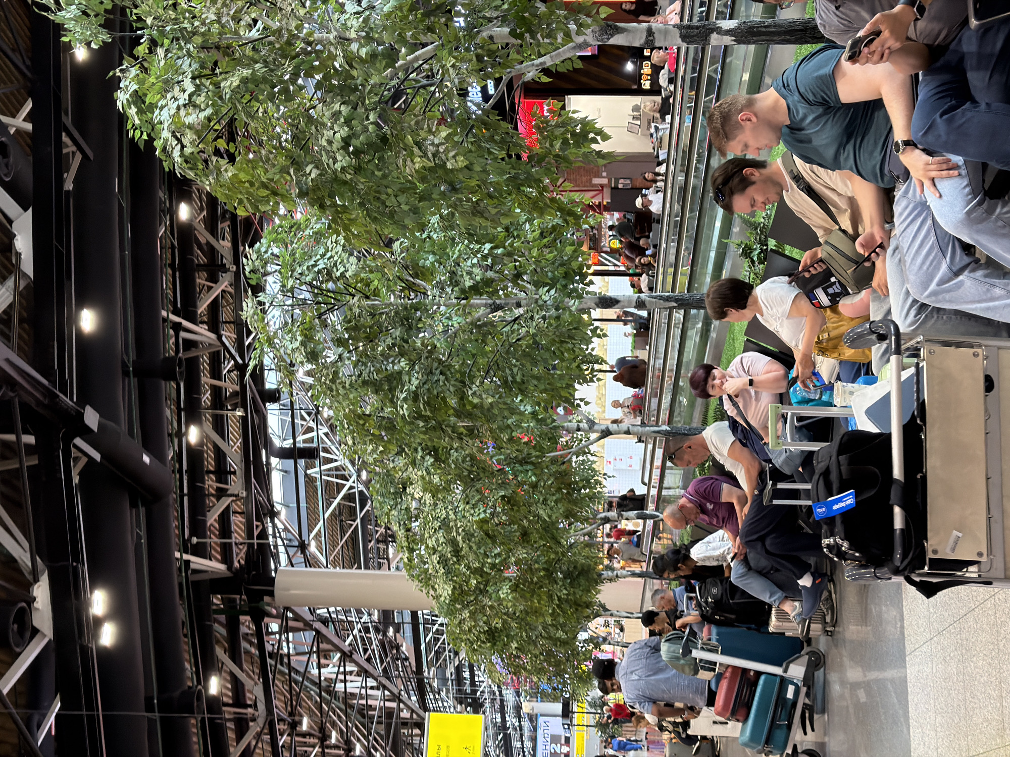 The image depicts a waiting area in an airport terminal, likely within the city of Khimki, Russia, as indicated by context clues. The scene is populated by a diverse group of people seated on black benches, many of them engaged with their mobile phones or conversing amongst themselves. Luggage, including suitcases and carry-on bags, is scattered around the seating area, some on trolleys. Overhead, the airport's infrastructure is visible, with exposed beams, ductwork, and lighting systems. In the foreground and mid-ground, artificial trees with green foliage are strategically placed, adding a touch of nature to the otherwise utilitarian environment. Some people are standing, while others are seated, appearing to be waiting for flights or to greet arrivals. The overall atmosphere suggests a typical airport scene characterized by transit and waiting.