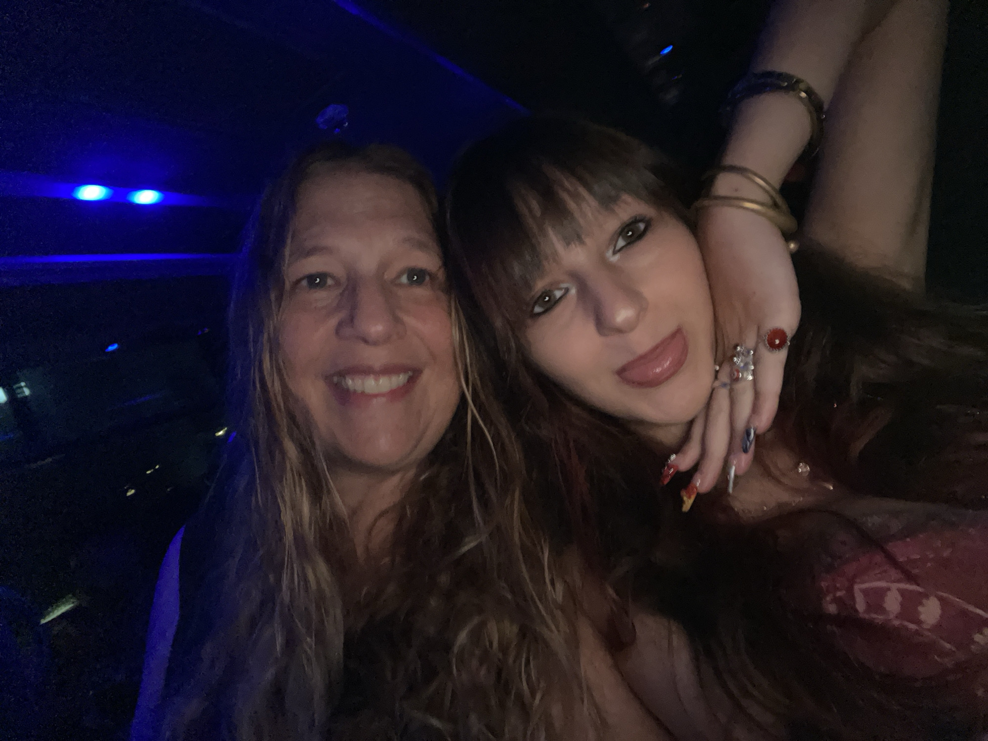 Two individuals, appearing to be a woman and a younger woman, are posing for a selfie in a dimly lit environment with blue lighting. The woman on the left has long, wavy blonde hair and is smiling. The younger woman on the right has dark brown hair with bangs and is sticking her tongue out. She is wearing several rings on her fingers and bracelets on her wrist. The background appears to be the interior of a vehicle or a room with dark walls and accents of blue light. The image does not contain any identifiable landmarks or features that would indicate a specific location in Herzliya, Israel.