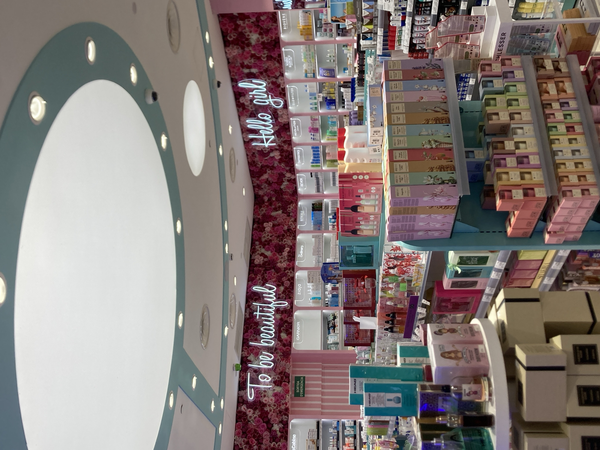 The image depicts the interior of a brightly lit retail store, likely a pharmacy or beauty shop, with a pink floral wallpaper accentuating the upper walls. Two prominent neon signs are visible: "To be beautiful" on the left and "Hello girl" on the right. The shelves are stocked with a variety of cosmetic and personal care products, including perfumes, lotions, and skincare items, organized in colorful packaging. The store has a modern and clean aesthetic with white shelving units and a light-colored ceiling. There are no people visible in the image, and the scene is static. The overall impression is one of a well-organized and visually appealing retail environment, possibly a chain store given the branding visible on some shelves. The mention of Barcelona, Spain, in the prompt suggests this is a shop located in that city.