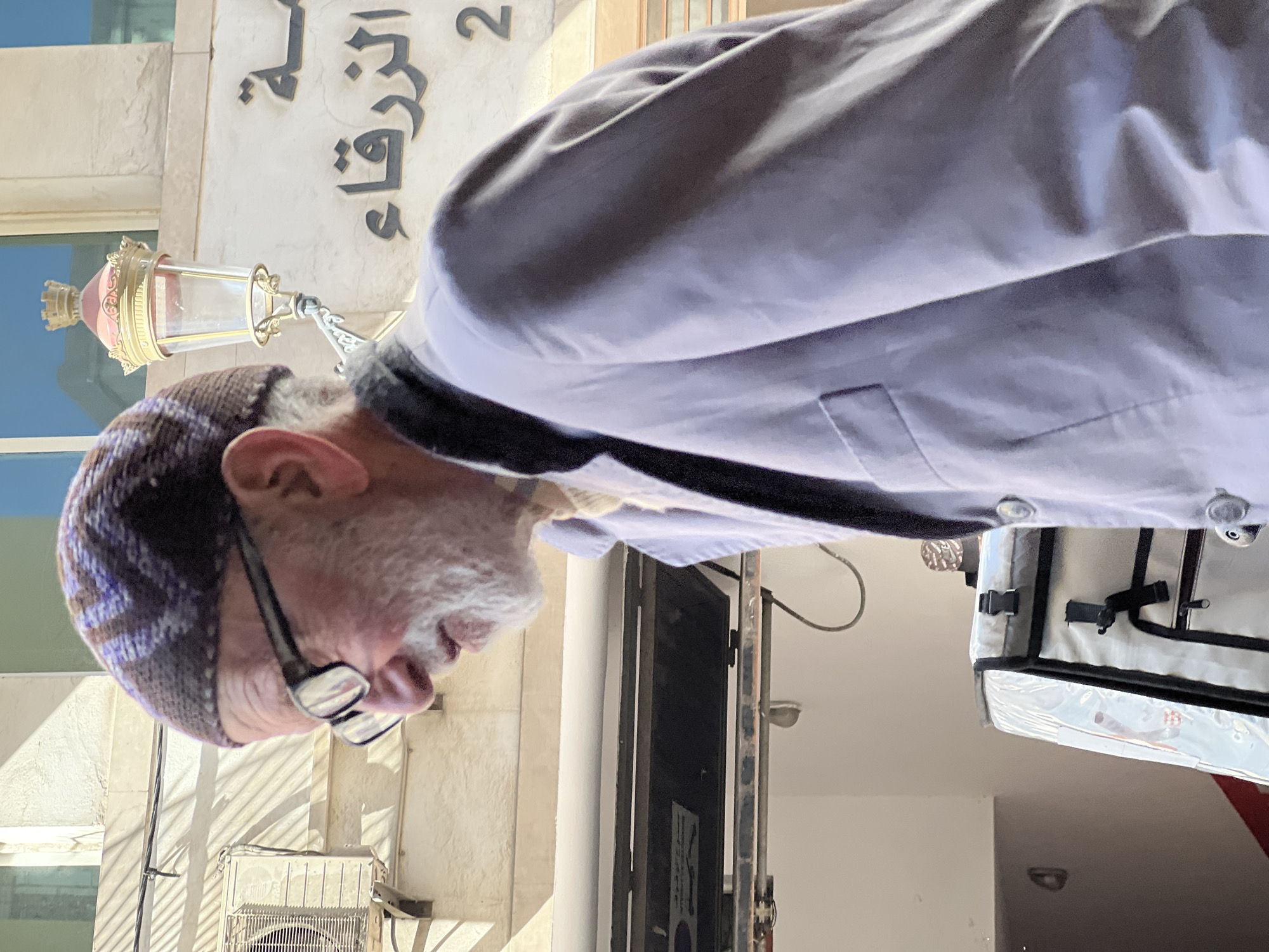 A man wearing glasses and a patterned skullcap is standing near a building with Arabic writing and the number 2 on its facade, potentially in Tanger (Tangier), Morocco. He's wearing a light-colored shirt or jacket. In the background, there is a lamp fixture attached to the building and what appears to be an air conditioning unit. Part of a delivery bag is also visible.