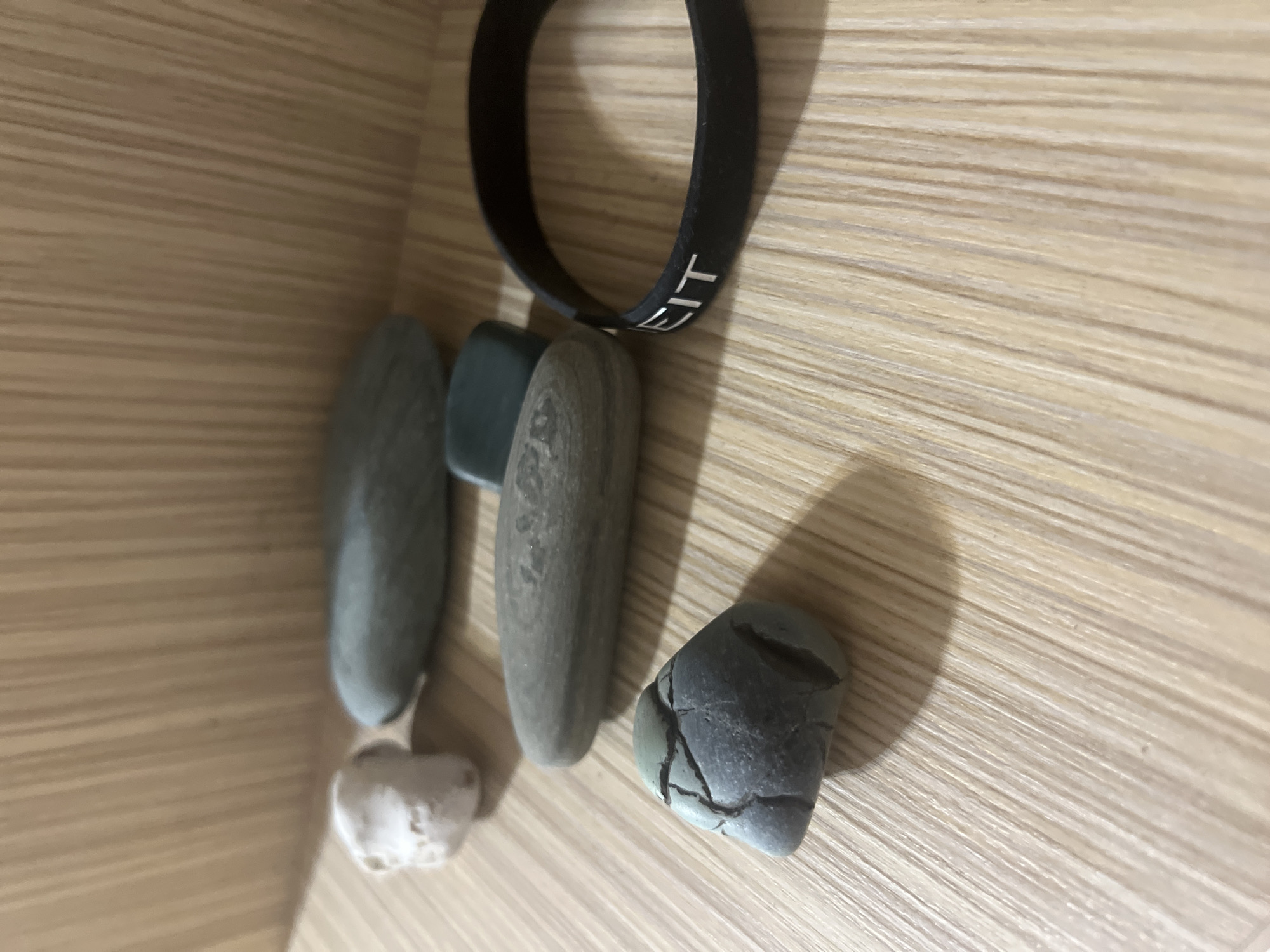 The image shows a close-up, eye-level shot of several objects placed on a light-colored wooden surface with distinct vertical grain. In the foreground, a small, dark gray, irregular-shaped rock with visible cracks sits on the left. To its right, a black silicone wristband with white lettering, partially visible as "EIT", is placed. Behind these objects, further to the left, are several smooth, gray, flattened stones of varying sizes. The largest of these is in the middle ground, oriented horizontally. To the left of this, and slightly further back, is a small, rough, white object. A dark green, rectangular object is also visible behind the middle gray stone. The background consists of vertical wooden planks, suggesting a shelf or wall. The lighting is soft, casting shadows from the objects onto the wooden surface. There is no indication of any specific location within the city of Surgut, Russia, or any human presence.