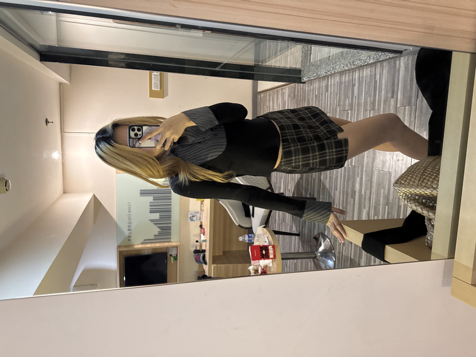 The image depicts a mirror selfie taken within a hotel room or similar private accommodation located in Yuzhong District, China.
The central subject is a young individual, likely female, with long blonde hair featuring visible dark roots, styled down. The person is wearing a long-sleeved black top with a grey and black striped high collar and matching cuffs, paired with a dark green and brown plaid mini skirt. A smartphone, held in the right hand with a light-colored, possibly marbled or patterned case, obscures the face as the individual captures their reflection. The left hand rests gently on the top edge of a light wooden bed frame or dresser in the foreground.
The environment, visible through the mirror's reflection, includes a light-colored wall with a large framed artwork featuring a barcode-like graphic and Chinese characters that read "我爱城市" (Wǒ ài chéngshì, meaning "I Love City"). Below this, a light wooden desk or vanity holds a dark flat-screen television on the wall above it, and various small objects on its surface, including a red baseball cap and cosmetic items. To the right of the desk, an upholstered light grey chair with dark legs is partially visible. A small round table with a metallic base sits in front of the chair, displaying a red box, consistent with a Chinese cigarette pack (e.g., Honghe brand), and a clear plastic water bottle. The floor is covered with grey wood-look laminate or tiles. To the far right, a tall, narrow wooden-framed glass partition or window is present. A woven-texture, light-colored handbag is seen on the floor next to the mirror in the lower foreground. The ceiling features recessed lighting fixtures.