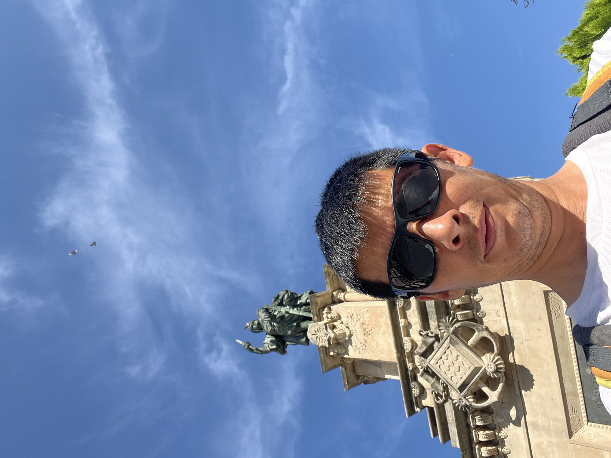 In Tarragona, Spain, a man in sunglasses takes a selfie with a clear blue sky and wispy clouds overhead. Behind him, a monument features a statue of a figure holding a sword aloft. The monument's facade displays intricate carvings, including what appears to be a shield or crest flanked by anchor-like motifs. The man is wearing a white t-shirt and a backpack. Two birds are visible flying in the upper portion of the sky.