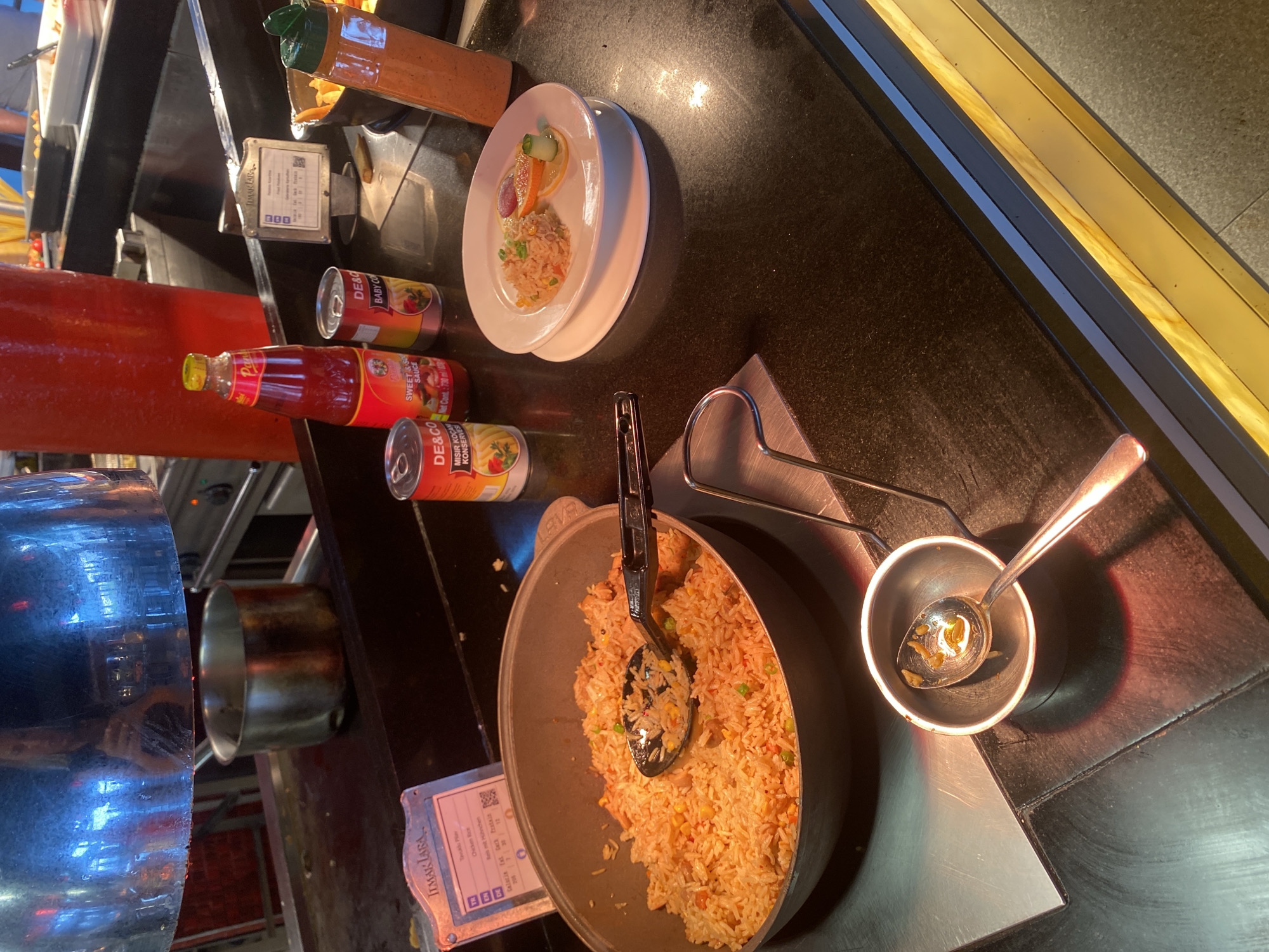 The image depicts a buffet station, likely in a restaurant or hotel, with various food items and condiments. In the foreground, a large metal pot filled with fried rice is visible, with a serving spoon resting inside. To the right, a white plate holds a small portion of fried rice garnished with slices of orange, lemon, and cucumber. Several bottles and cans of sauces and condiments are arranged on the counter behind the plate, including a bottle of Sriracha-like sauce, and cans of DE&CO baby corn and sweet & sour sauce. A metal container holding a used spoon is positioned in the lower left corner. In the background, a large red container and a metal pot under a heat lamp suggest a self-serve food area. The lighting is warm and focused on the food items. The environment appears to be an indoor dining area with a dark countertop and reflective surfaces. There are no people clearly visible in the image. The sign in the upper left corner of the image appears to have text in Turkish and German, indicating a location in Turkey. Based on the Turkish text, the location is identified as Aksu, Turkey.
