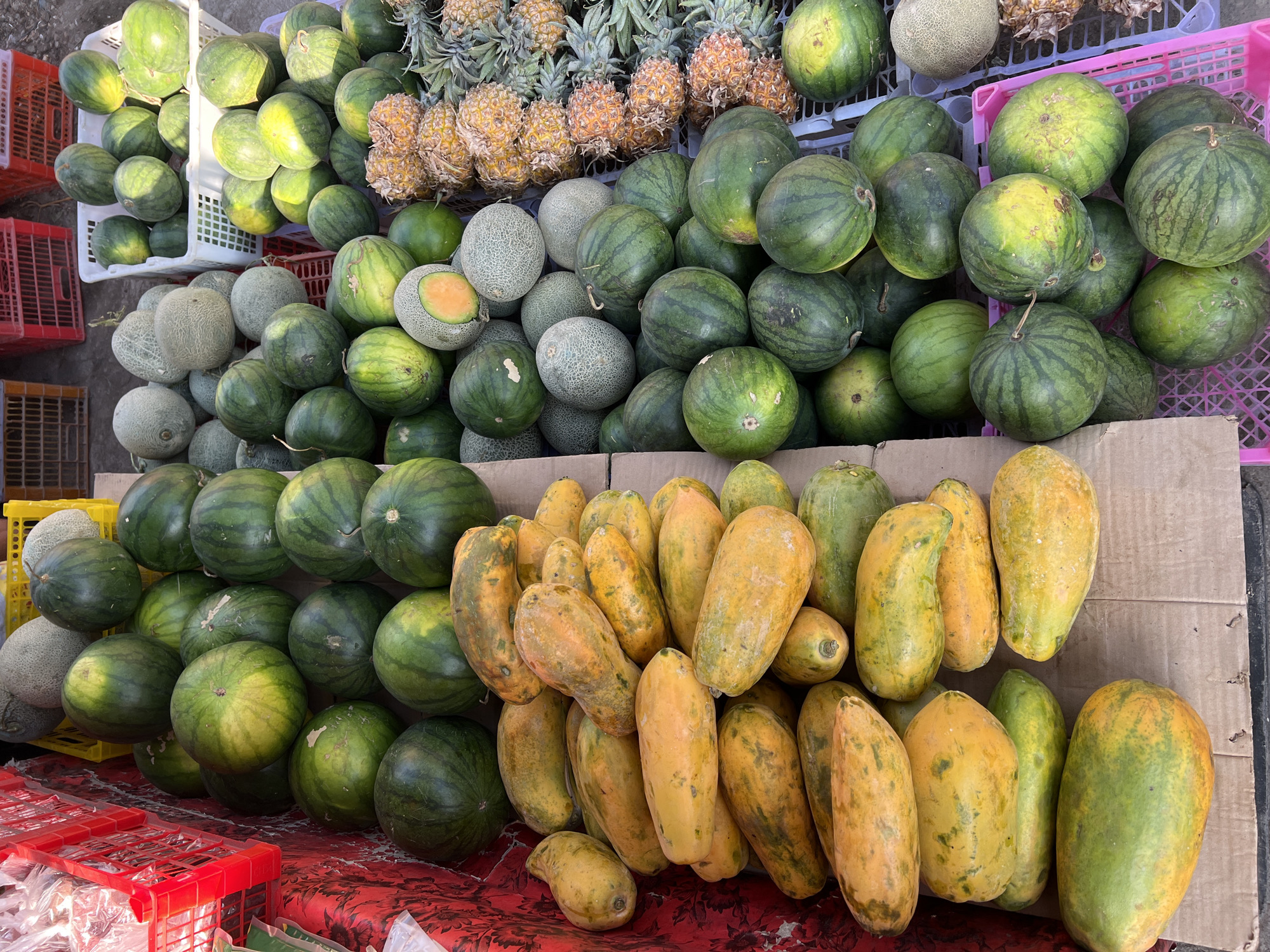 This is a high-angle, full shot of a fruit stall in the city of Ubud District, Indonesia. The stall is filled with a variety of fruits, including watermelons, cantaloupes, papayas, and pineapples. The fruits are arranged in baskets and on cardboard. The stall appears to be outdoors, possibly on a street or market. The lighting suggests it is daytime. There are no people visible in the frame.