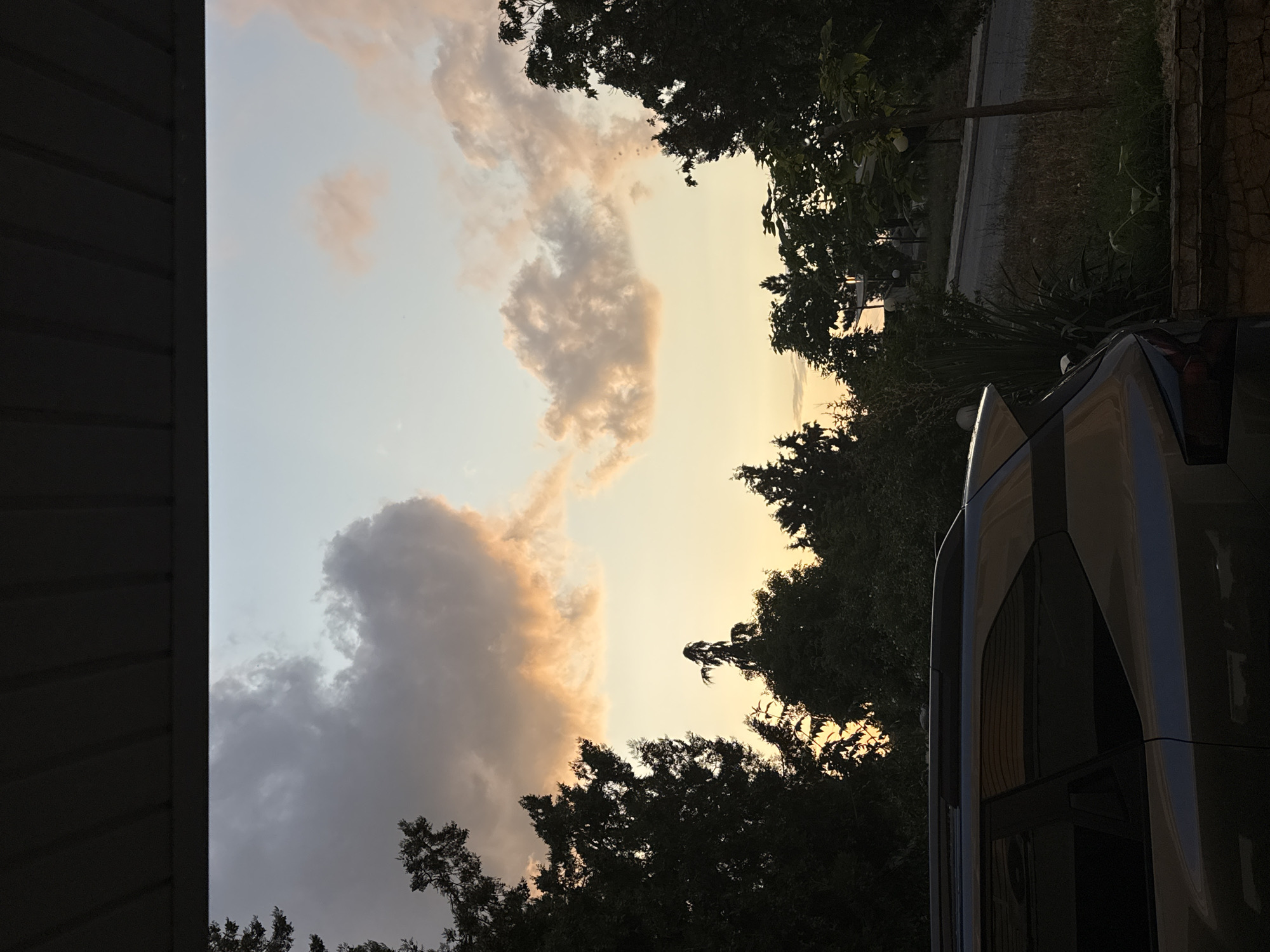 The image shows a view from under a dark, corrugated overhang, looking out at a dusky sky. Silhouetted trees, some tall and slender, others dense and bushy, frame the lower part of the sky. The sky itself transitions from a pale blue at the top to a warm, peachy orange near the horizon, illuminated by the setting sun. Scattered clouds, tinged with pink and grey, drift across the sky. In the foreground, the rear half of a silver SUV is visible, its windows dark and reflective. To the right of the car, a low stone wall runs horizontally, bordering overgrown grass and a narrow, winding road. A slender tree with a few small, light-colored spheres hangs from its branches stands near the road. The scene appears to be an outdoor residential area in Sredets, Bulgaria, possibly a driveway or a patio area, during sunset.