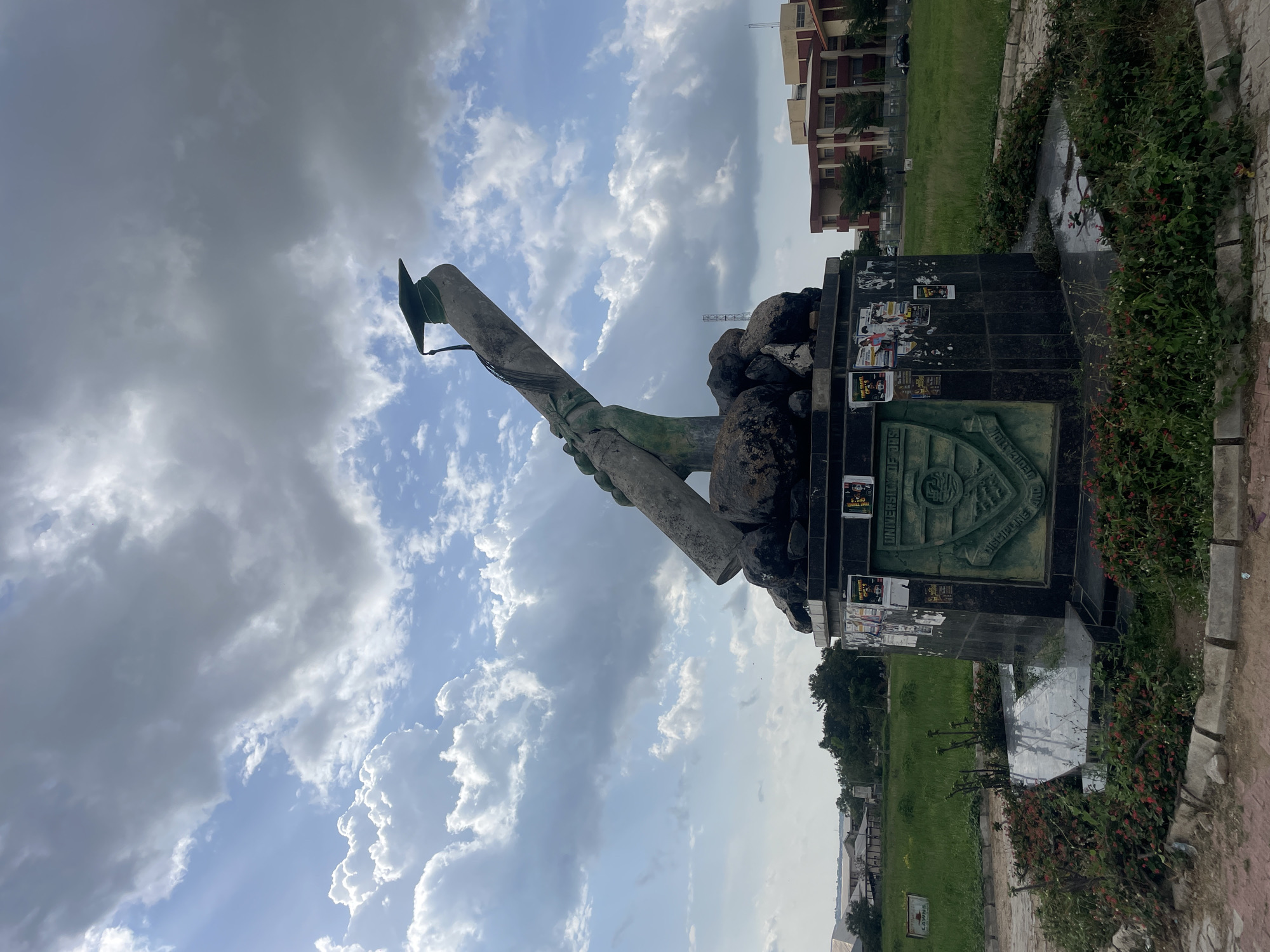 The image displays a monument set against a partially cloudy sky, depicting a sculpture of two hands holding a rolled scroll, crowned with a graduation cap. The sculpture, crafted from a weathered grey material, likely concrete or stone, shows signs of green patination. It rests on a dark, polished base, possibly made of granite or marble.
On the front of the base, an embossed circular seal is visible, featuring partially legible text including "UNIVERSITY OF..." and "...DISCIPLINE AND...". Numerous paper flyers and posters are haphazardly pasted onto the dark base, with phrases such as "THE TRAVELLERS" and "UNIVERSITY" discernible on some. Behind the main sculpture, on top of the base, are several large, rough dark rocks.
The monument is situated within a green, grassy expanse. Around the base, there are garden beds with small flowering plants, displaying both red and white blossoms. In the background, a multi-story building with reddish-brown walls and lighter upper sections is visible, suggesting an institutional setting. Further in the distance, other structures and green areas can be seen.
The sky occupies a significant portion of the image, showcasing a mix of bright white cumulus clouds and darker grey clouds, indicating a dynamic weather pattern typical of daytime. No people are visible, and no specific activity is taking place. The academic theme of the monument, featuring a graduation cap and scroll, strongly suggests that the location is a university campus or an educational institution, consistent with the visible text mentioning "UNIVERSITY." The overall impression is a mid-day scene under varied cloud cover.
