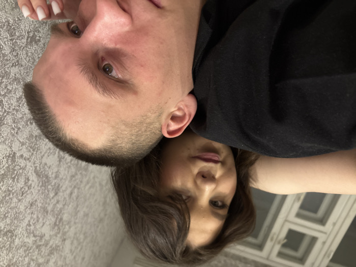 A man and a woman are close together in a photo taken from a low angle. The man has a shaved head and looks directly at the camera with focused eyes. He is wearing a black shirt. The woman is behind him, looking at the camera with a soft expression. A white window is visible in the background on the right. The entire scene is slightly rotated counterclockwise.