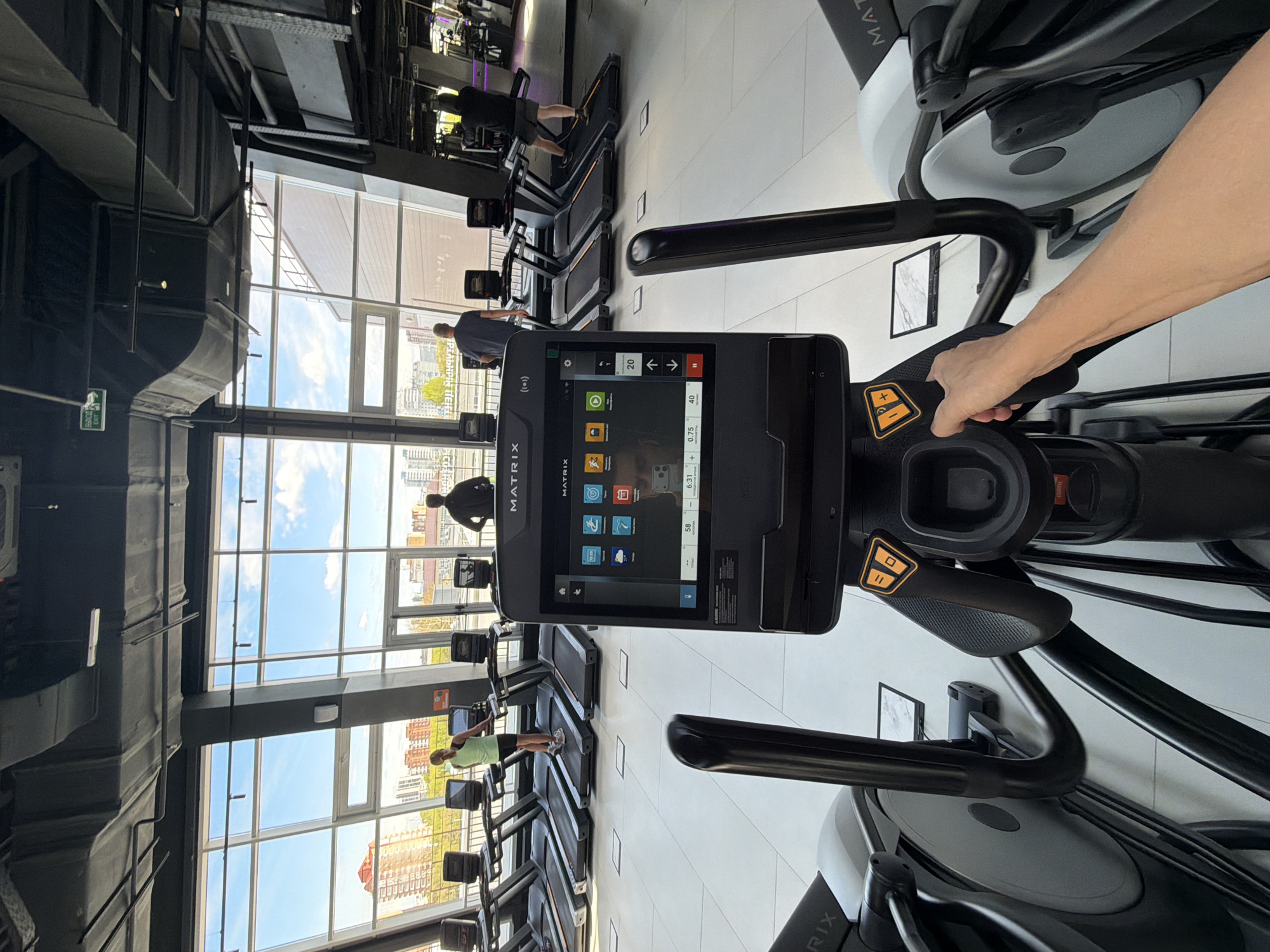 The image depicts the interior of a gymnasium or fitness center in Krasnoyarsk, Russia, as evidenced by the buildings visible through the large windows. The focus is on a Matrix-branded treadmill or elliptical machine, with a digital display showing various functions and workout data. A person's hand is visible on the control handles of the machine, interacting with the controls. The gymnasium features multiple treadmills arranged in rows, facing a large window with city views. Several individuals are present, using the exercise equipment. The overall environment is modern and well-lit, with visible ceiling fixtures and structural elements.