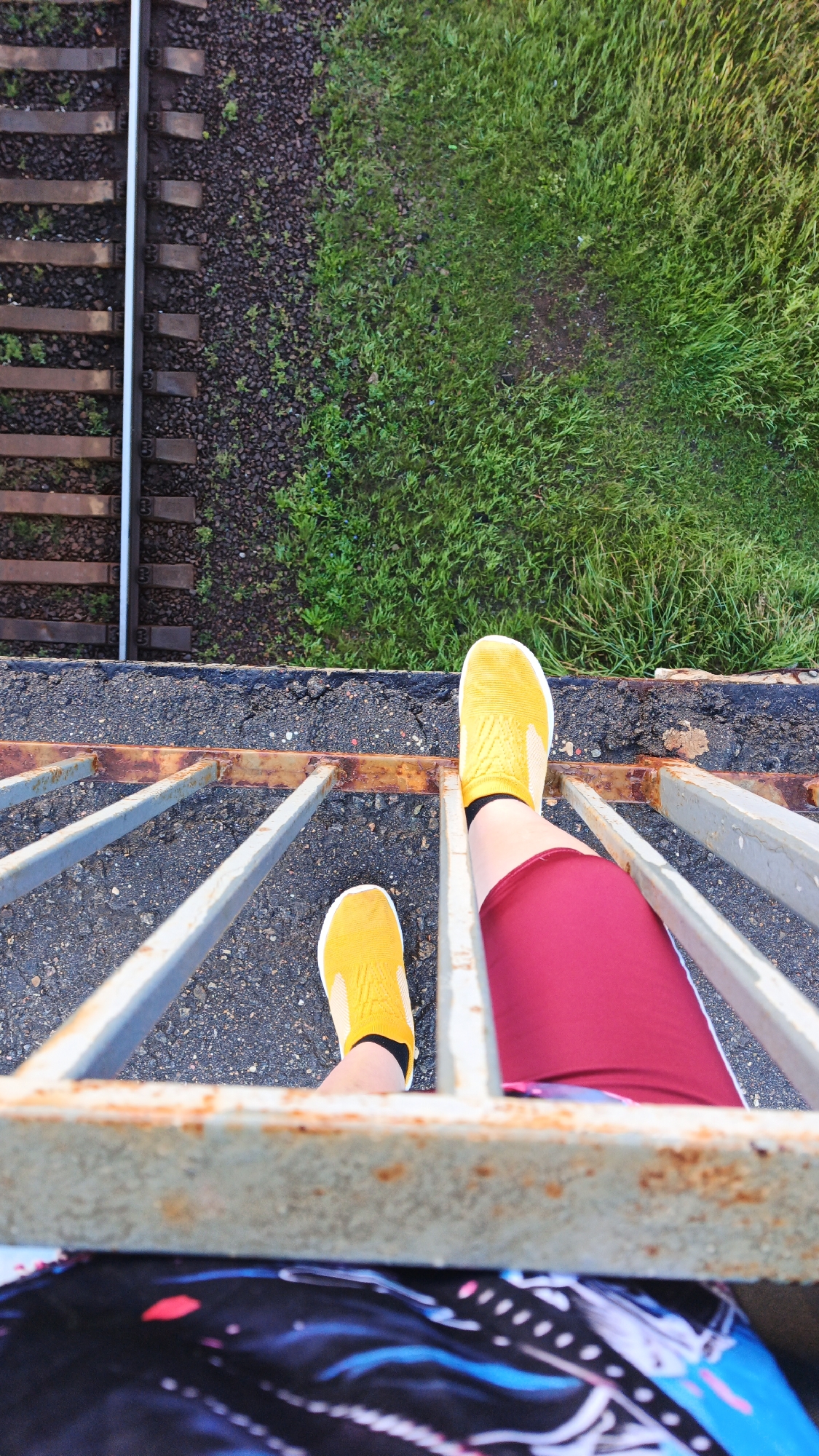 This is a high-angle, vertical shot looking down at a person's legs and feet, standing on a rusted metal railing. The person is wearing yellow slip-on sneakers and dark red leggings. Below the railing, a section of patterned fabric is visible, possibly part of clothing. The railing overlooks a railway track to the left and a grassy area to the right. The railway track consists of wooden sleepers and ballast. The grass appears somewhat overgrown. The overall environment suggests an outdoor location, possibly an overpass or bridge near a railway line in Drezna, Russia. The lighting suggests it is daytime.