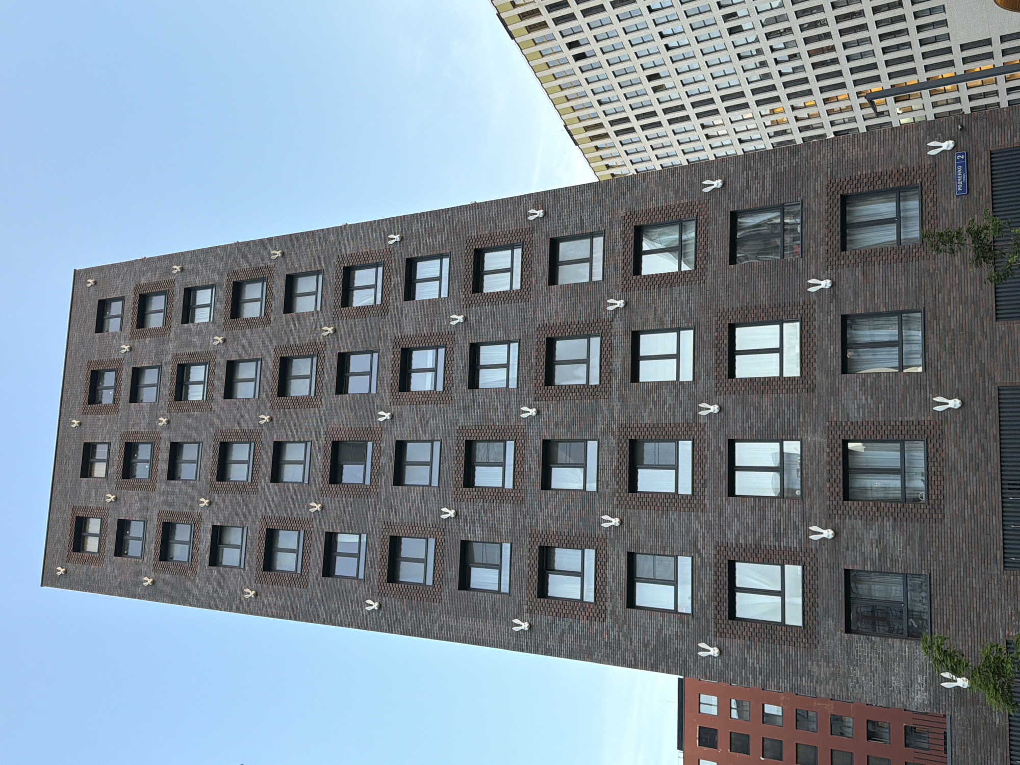 The image captures an upward view of a modern brick apartment building in Moscow, Russia, characterized by its dark brown brick facade and numerous windows. The building is adorned with white rabbit sculptures placed between the windows. To the right of this building, another contemporary apartment building with a white facade and a patterned grid of windows is partially visible. The sky above is clear and pale blue. A street sign that reads "РОДЧЕНКО 2" is visible at the lower right of the frame. No people are present in the image.
