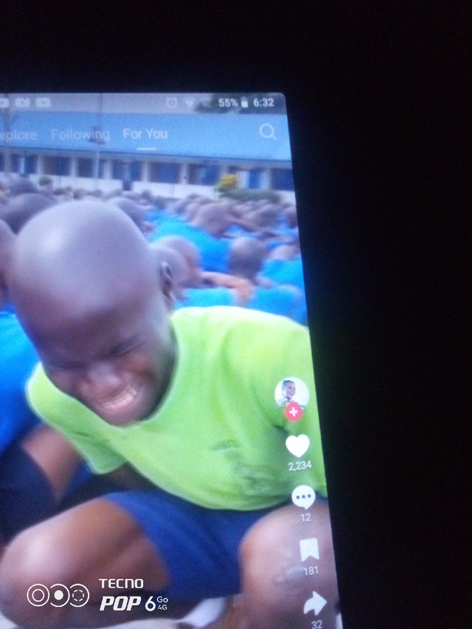 The image displays a close-up of a person with a shaved head wearing a bright green shirt and blue shorts. They are looking down and appear to be laughing or smiling broadly, with their eyes closed. In the background, a group of people, likely in uniform, are visible. The image is a screenshot of a mobile phone screen, with elements of a social media interface such as likes, comments, and shares displayed on the right side. The phone's battery and time are visible at the top. The phone is a TECNO POP 6 4G. There is no indication of the location being Howayi, Nigeria, or any specific city.