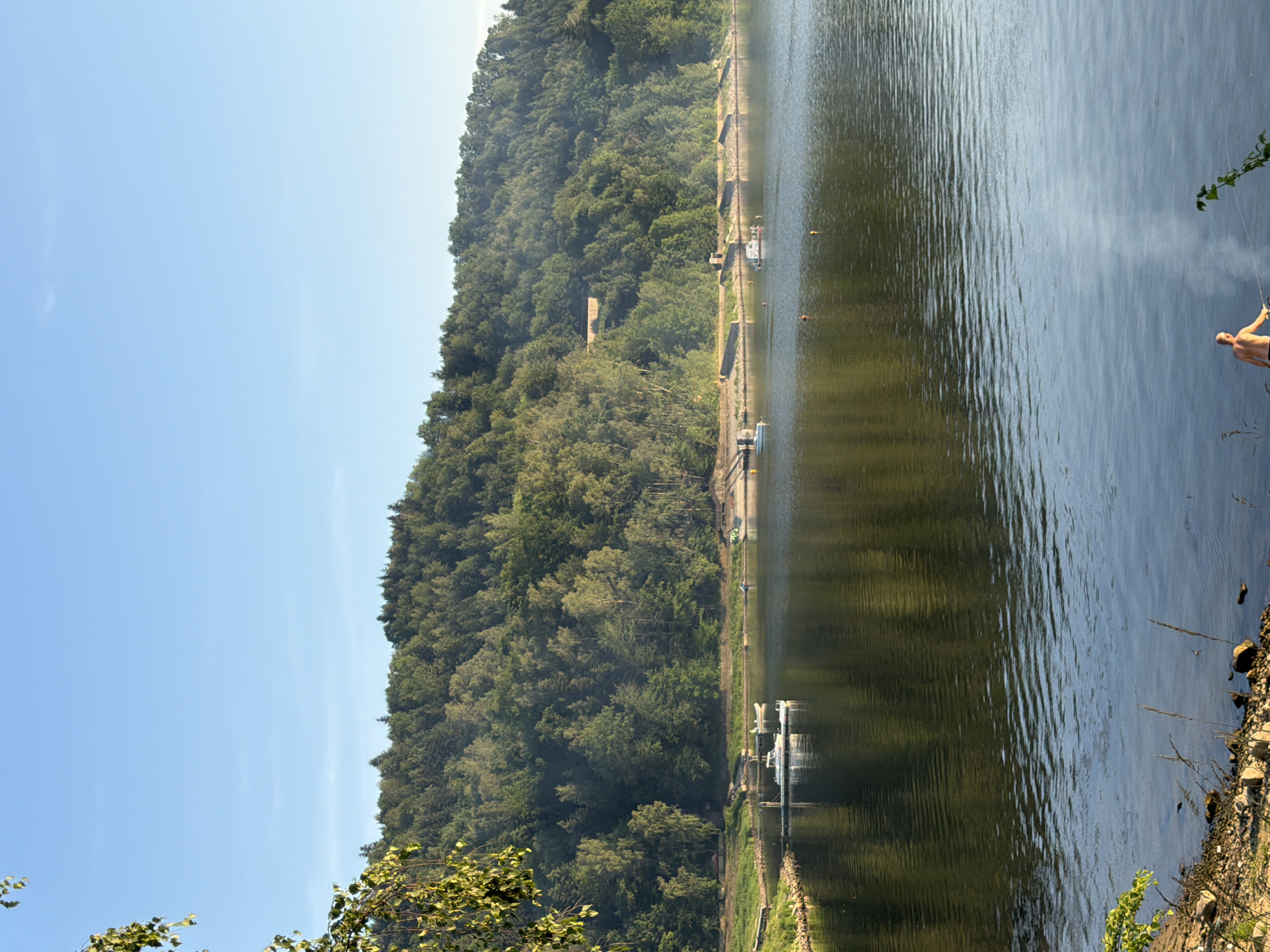 The image shows a body of water with a forested hill in the background. There are several boats on the water, including a white boat docked at a pier. A person is visible on the shore in the foreground, holding a fishing rod. The sky is clear and blue. The scene is likely Lipie, Poland, based on the provided information.