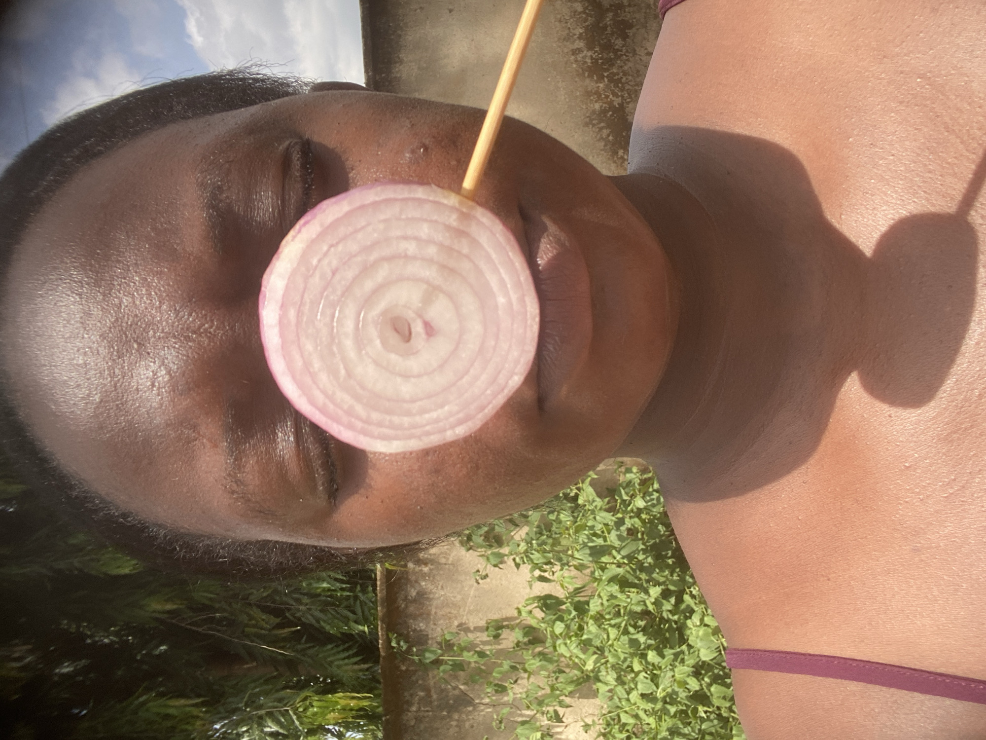 An outdoor, close-up, and slightly overhead shot features a person with dark skin, seen from the upper chest to the top of the head, against a bright background. The person's eyes are closed. A circular slice of red onion is positioned directly in front of their mouth and nose area, held in place by a thin wooden skewer or stick that extends upwards from behind the onion.
The scene is brightly lit by direct sunlight, casting strong shadows on the person's skin, particularly on their right shoulder and chest. The person appears to be wearing a dark reddish or maroon strap, possibly from a top or swimwear, visible on the lower right.
The background on the left side shows a clear blue sky with some white clouds. On the lower right and bottom, green foliage and plants are visible, suggesting a natural outdoor setting. A concrete or light-colored wall or structure is also discernible in the upper right background. The image's brightness and sharp shadows indicate it was taken during daylight hours, likely midday or early afternoon, in Maigizo, Nigeria. There is no visible text in the image.