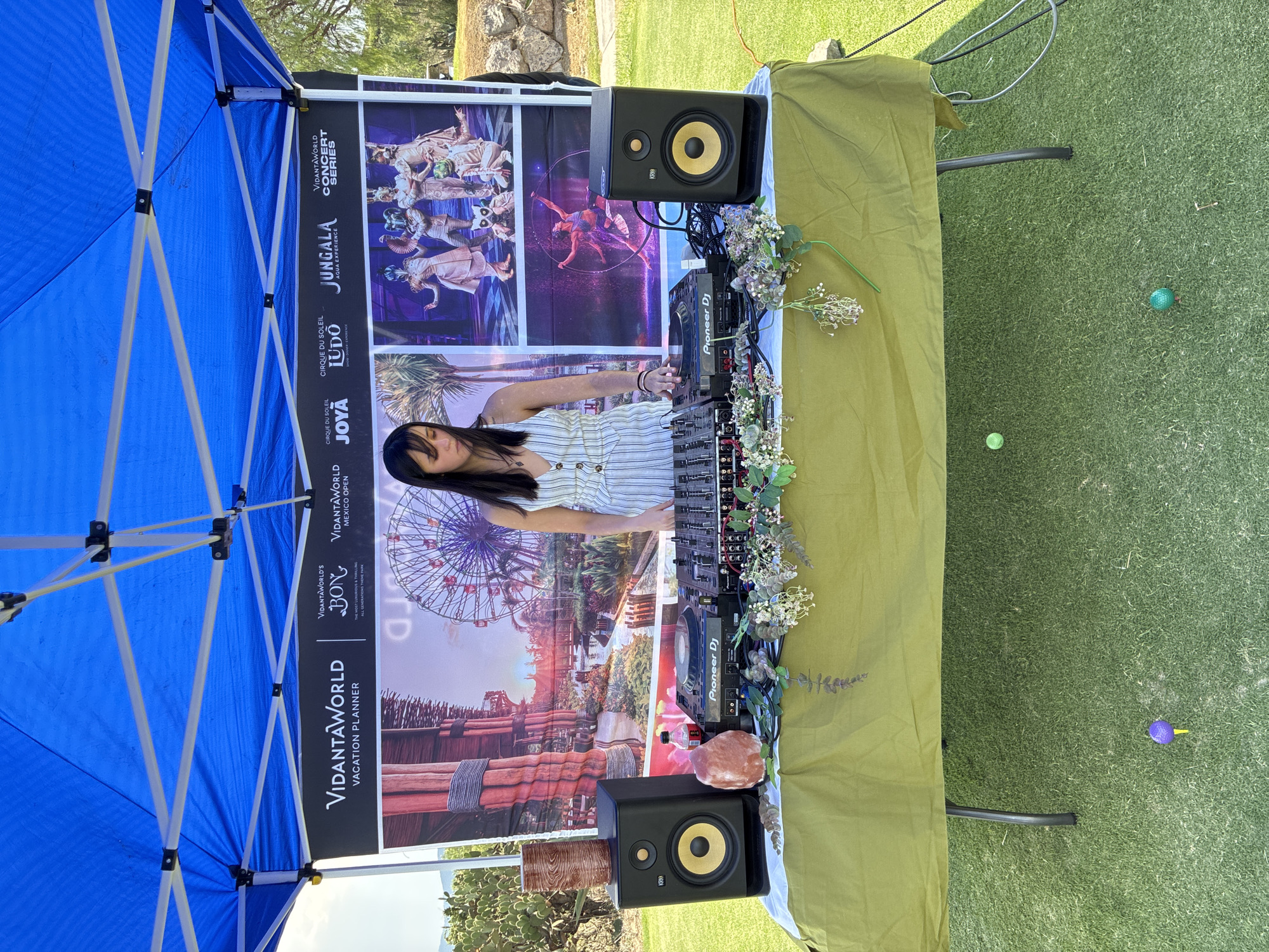 A vertical, outdoor shot shows a DJ performing in Pueblo Nuevo de Jasso, Mexico. The DJ is a young woman wearing a white-striped shirt. She is standing behind a Pioneer DJ setup on a table covered with a green tablecloth, which is decorated with flowers. The DJ is adjusting the controls on the mixer. Behind the DJ setup is a large banner with the logos of various brands, including VidantaWorld, Cirque du Soleil, and Jungala. The banner features images of the attractions offered by these brands. The DJ setup is flanked by two KRK speakers. The DJ is under a blue tent. The ground is covered in artificial turf. Several golf balls can be seen on the turf.