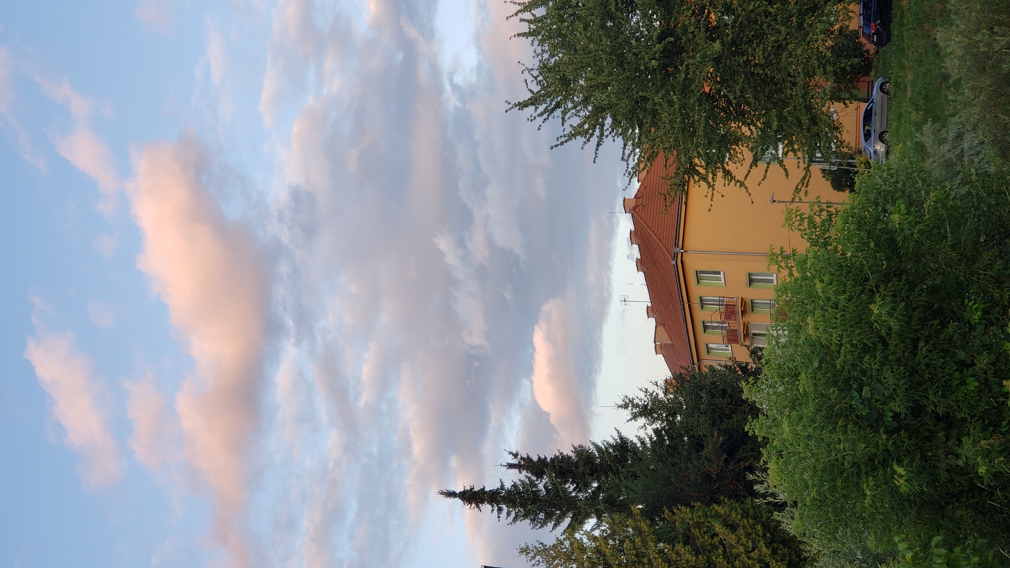 The image depicts a residential building in Prešov, Slovakia, under a cloudy sky tinged with the soft hues of sunset. The building, with its orange facade and red tiled roof, features multiple stories with balconies and windows. Evergreen trees and lush green bushes surround the structure, and a few cars are parked in the grassy area in front of it. The scene is captured in a vertical orientation, emphasizing the vastness of the sky above the urban landscape.