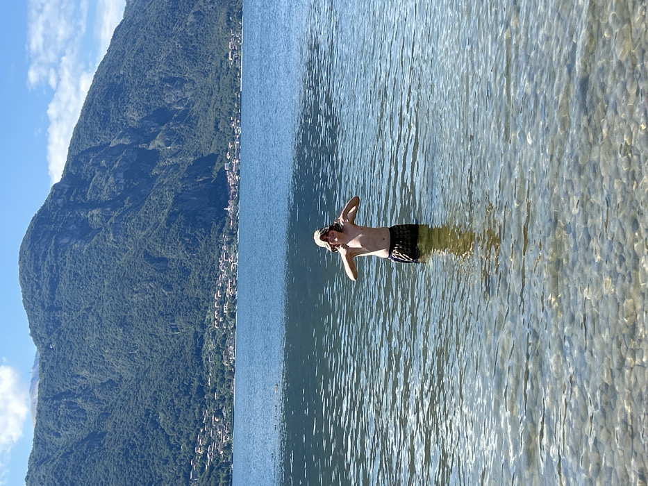 The image displays a male youth standing chest-deep in the clear, rippled waters of Lake Como, specifically in the area of Abbadia Lariana, Italy. The lakebed, composed of numerous rounded stones, is visible through the transparent water. The individual, shirtless and wearing dark, patterned swim trunks and a light-colored bucket hat, faces forward with a visible smile. His arms are bent at the elbows, and both thumbs are pointed downwards. In the background, across the expanse of the lake, a series of densely forested mountains rise, with a village or small town composed of light-colored buildings with red roofs nestled along the distant shoreline at their base. The sky above is bright blue with scattered white clouds.