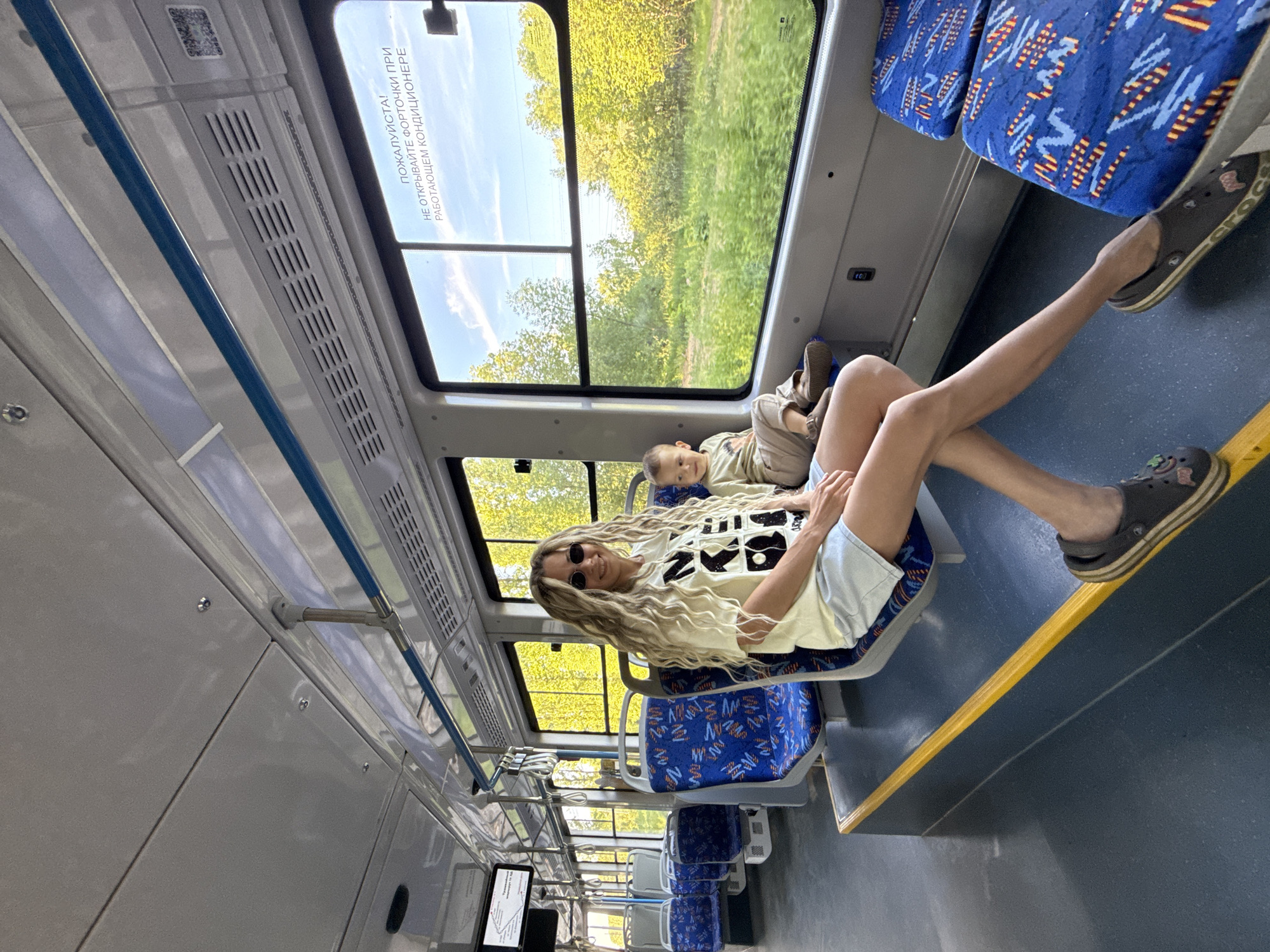 Inside a public transport vehicle, likely a tram or bus, in the city of Шебунино, Russia, a woman and a young child are seated on the right side. The vehicle is in motion, evidenced by the slight blur of the lush green landscape outside, visible through large windows under a bright, partly cloudy sky.
The adult female, positioned closer to the camera, has long, wavy blonde hair and wears dark oval sunglasses, a light-colored short-sleeved t-shirt with a black graphic, and light denim shorts. Her legs are extended, with her feet, clad in dark Crocs-style shoes with striped soles, resting on a yellow-trimmed elevated floor section. She looks towards the camera with a subtle smile.
To her right, a young child with light hair is seated, looking towards the window. He wears a light green long-sleeved shirt with a graphic and light brown trousers, along with dark slip-on shoes.
The interior features multiple rows of blue seats with a geometric pattern, grey frames, and dark grey-blue speckled flooring. A prominent yellow safety line marks the edge of the elevated seating area. Overhead, a white ceiling holds blue horizontal grab rails and multiple air vents. Russian text on the window advises, "PLEASE! DO NOT OPEN WINDOW VENTS WHILE AIR CONDITIONER IS RUNNING." A QR code is visible on the wall near the window. In the upper left background, a digital display screen shows what appears to be a route map with Russian text.