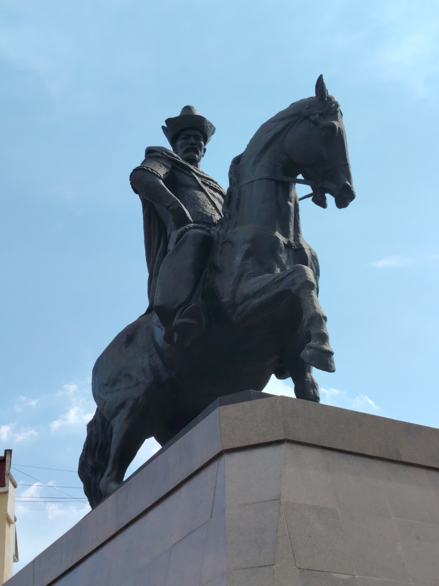 The image shows a low-angle shot of a dark bronze equestrian statue against a clear blue sky with scattered white clouds. The statue depicts a man in historical attire, including a wide-brimmed hat and a cape, seated on a horse. The horse is in mid-stride, with its front legs raised, giving a sense of dynamism to the sculpture. The base of the statue is made of light gray stone, with a rough, faceted surface. In the background, a portion of a building with a red-tiled roof and light-colored walls is visible to the left, along with thin wires stretched across the sky. The statue is located in Astana, Kazakhstan, and is known as the Kenesary Khan monument, dedicated to the leader of the Kazakh liberation movement.