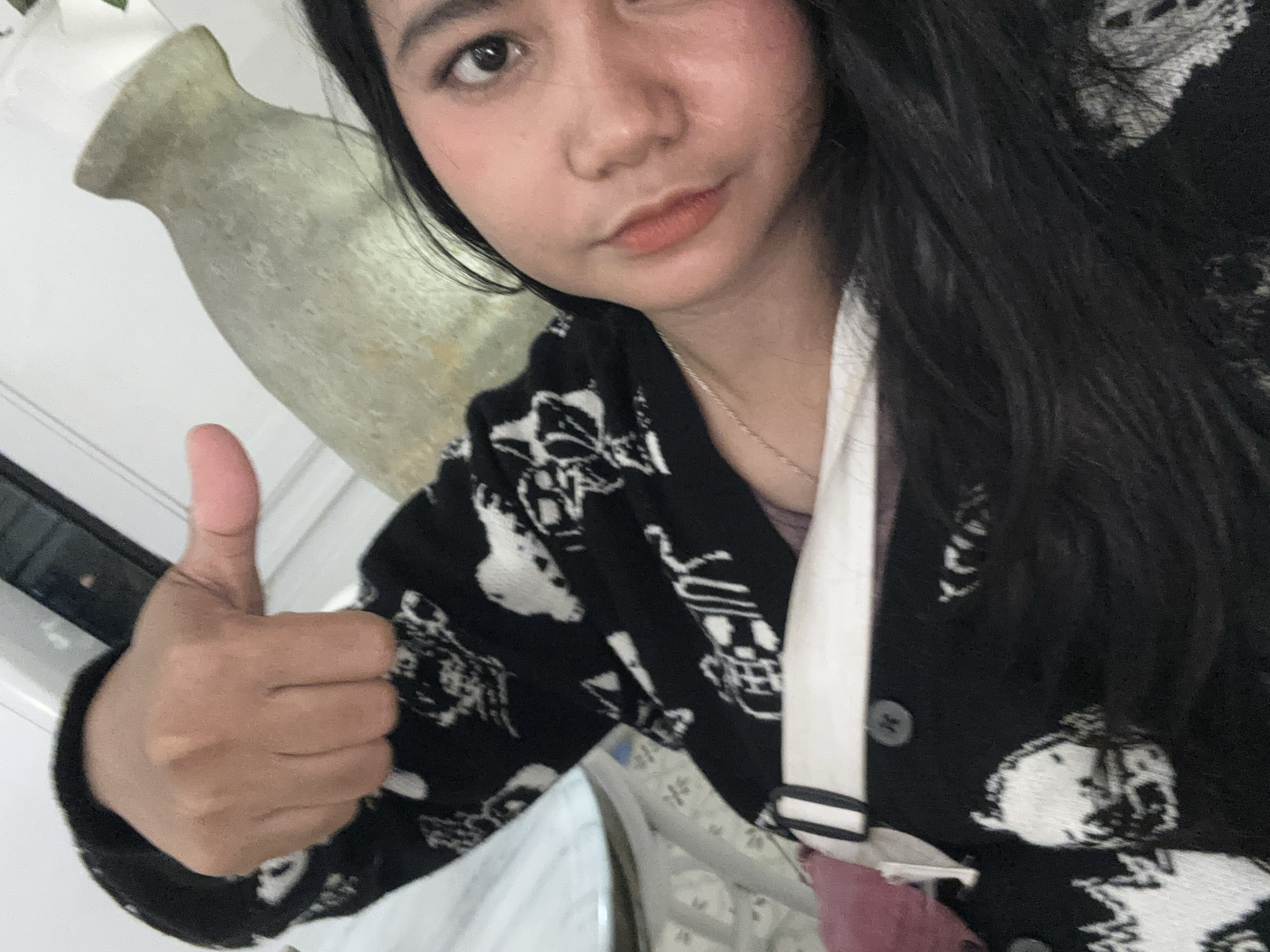 A young woman with long, dark hair and makeup is lying down and taking a selfie. She is wearing a black and white patterned cardigan over a white top. Her left hand is raised, giving a thumbs-up gesture. In the background, a light-colored, elongated vase or urn is visible. The scene appears to be indoors, with a white wall and furniture elements suggesting a domestic or possibly a cafe setting in West Jakarta, Indonesia.