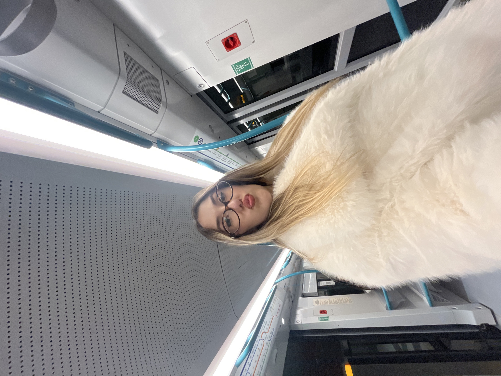 This image is a brightly lit, rotated selfie taken by a young woman inside a modern public transport vehicle, likely a metro or train in Sofia, Bulgaria. The photograph is oriented sideways, giving the impression that the ceiling is to the left and right.
The primary subject is a young woman with long, straight blonde hair, wearing round glasses and a fluffy white faux fur coat or jacket. She is looking directly at the camera with a playful expression, making a "kissing face" with puckered lips.
The setting is a clean, contemporary vehicle interior characterized by light grey and white panels. A long, bright LED light strip runs along the ceiling, providing ample illumination. A light blue, curved handrail extends through the upper part of the frame. On the ceiling, there is a prominent red emergency stop button housed in a white square panel. Below it, a green pictogram depicts a person with an arrow, possibly indicating an emergency exit or information point. Further along the ceiling, a grille, likely for ventilation or a speaker, is visible. To the left, a grey wall panel features a repetitive pattern of small circular perforations. Reflections in overhead panels suggest windows or other reflective surfaces. A metro map, displaying various colored lines and station names, is partially visible on the ceiling, typical of an urban transport system. A small green and white sticker with the letter "C" and another symbol is also present near the blue handrail. The overall impression is one of a comfortable and well-maintained public transport environment.