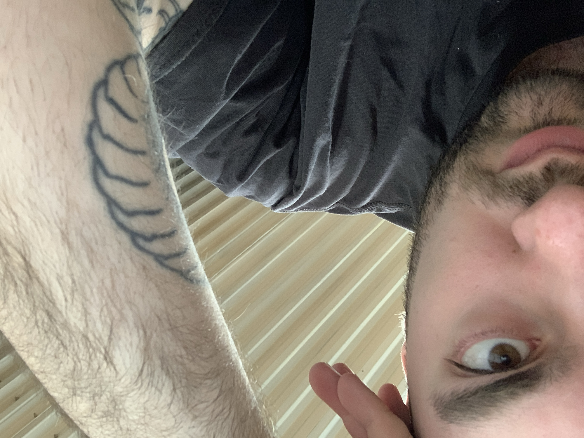 A close-up, low-angle selfie shows a person's face, arm, and upper torso. The person has brown eyes, a beard, and appears to be wearing a dark-colored shirt. The arm, with visible hair and a tattoo resembling a shell or stylized feather, is positioned to the left of the frame. The background consists of a light-colored, striped surface. The photo was not taken in Chaikovsky, Russia; there are no indicators to suggest this location.