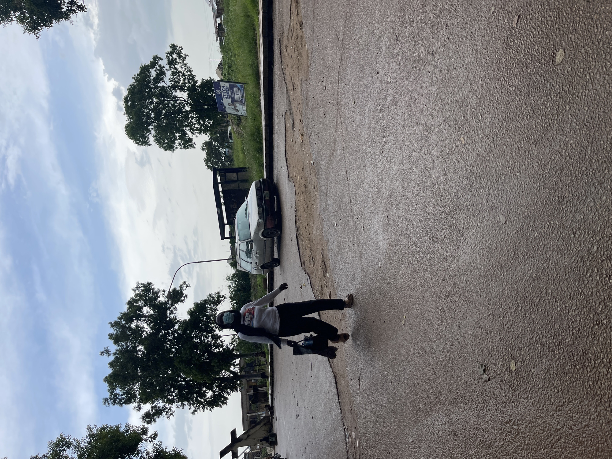 A person wearing a light-colored long-sleeved shirt, black pants, and a face mask is shown in motion, seemingly practicing dance or freestyle moves on a wet asphalt road. They are holding a dark-colored bag. The scene is set in Rafin Par, Nigeria, under a cloudy sky suggesting it might be overcast or after rain. Mature trees with dense foliage line the background, with one large tree prominently featured. A white car is parked near a damaged roadside curb. A street lamp is visible, and a sign with text that includes "STITCH" and an image is posted behind a tree. The overall atmosphere appears casual and the person's pose suggests an active, perhaps artistic or recreational, moment.