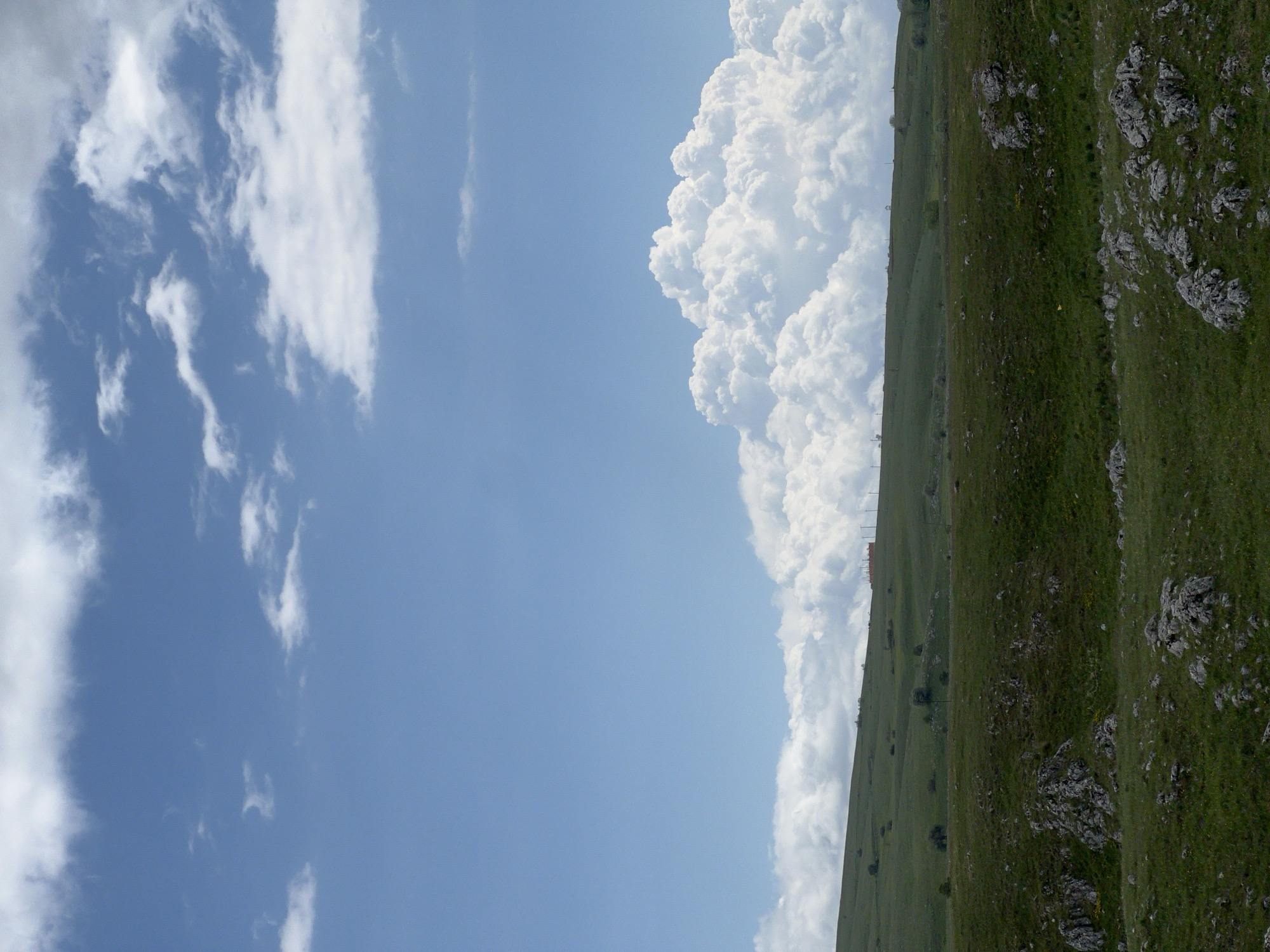 The image shows a view in Pišče, Montenegro. A grassy, rocky hillside occupies the right side of the frame. Above the hillside, a large cumulonimbus cloud formation rises into the sky. The left side of the image shows a blue sky with scattered cirrus clouds.